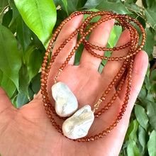 Load image into Gallery viewer, Hessonite Garnet and Baroque Pearl Lariat Necklace held in hand against green leaf backdrop, handcrafted in Canada.