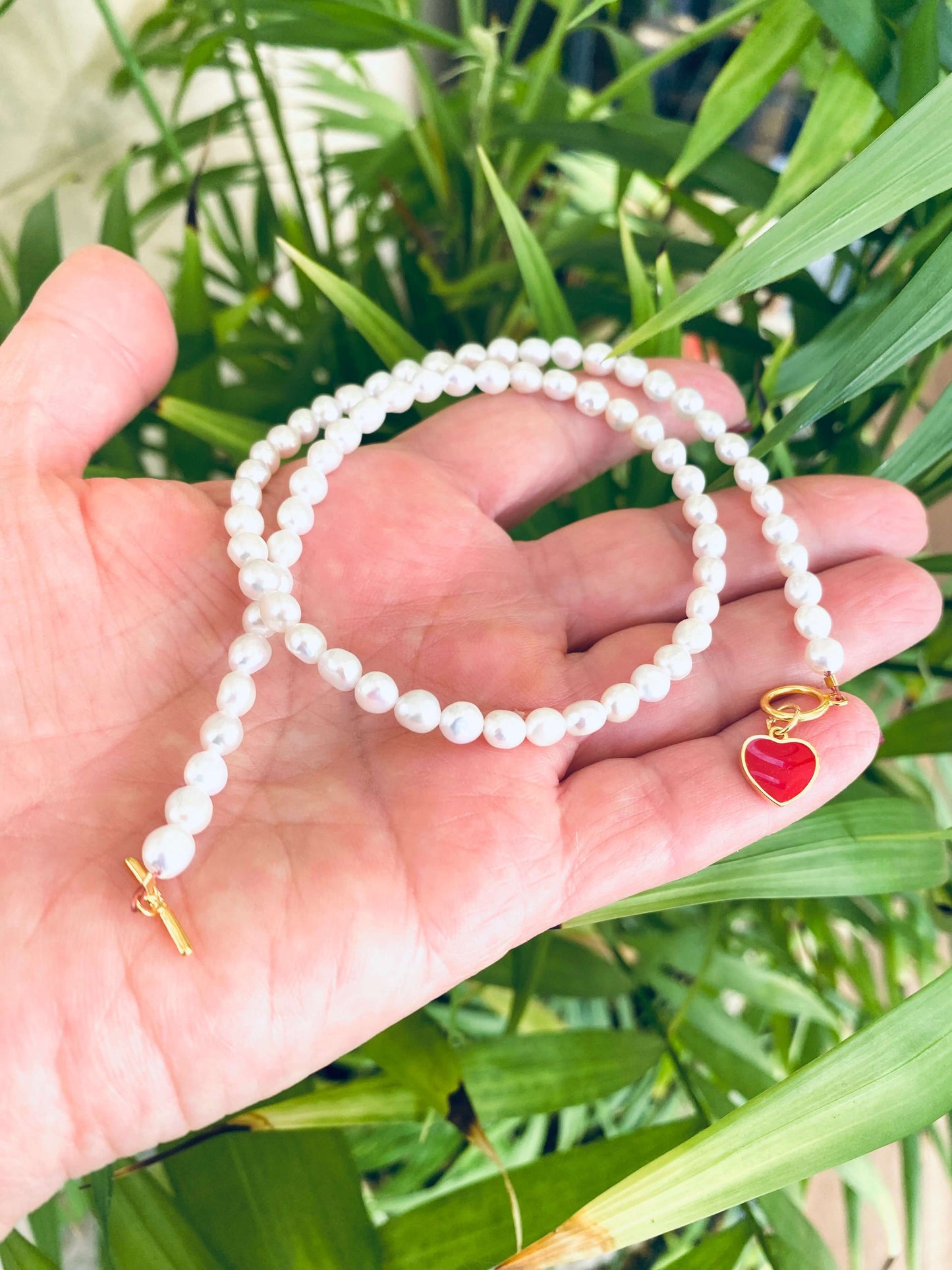 Handcrafted White Pearl Necklace featuring red heart charm and gold clasp held near green leaf background.