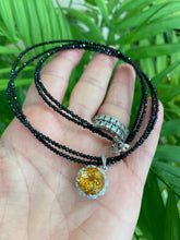 Charger l'image dans la galerie, Hand holding black spinel and citrine pendant necklace near green leaf background