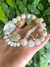 Charger l'image dans la galerie, Hand holding morganite and jade necklace with baroque pearls against green natural foliage, showing gemstone color and luster.