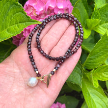 Load image into Gallery viewer, Garnet toggle necklace with baroque pearl charm displayed on hand among vibrant green leaves and pink flowers.