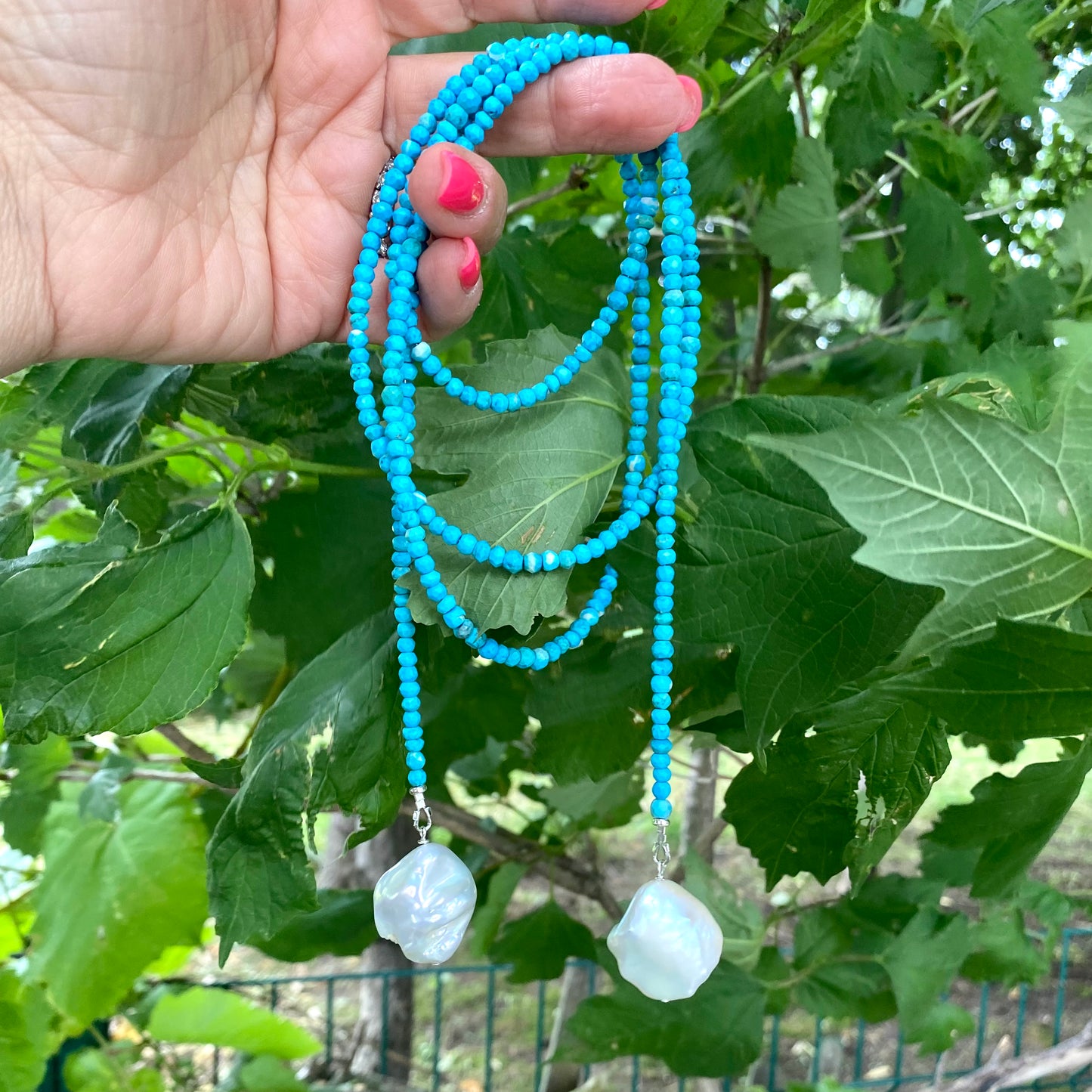 Turquoise lariat necklace featuring two baroque pearls on a vibrant turquoise bead strand held against green foliage.