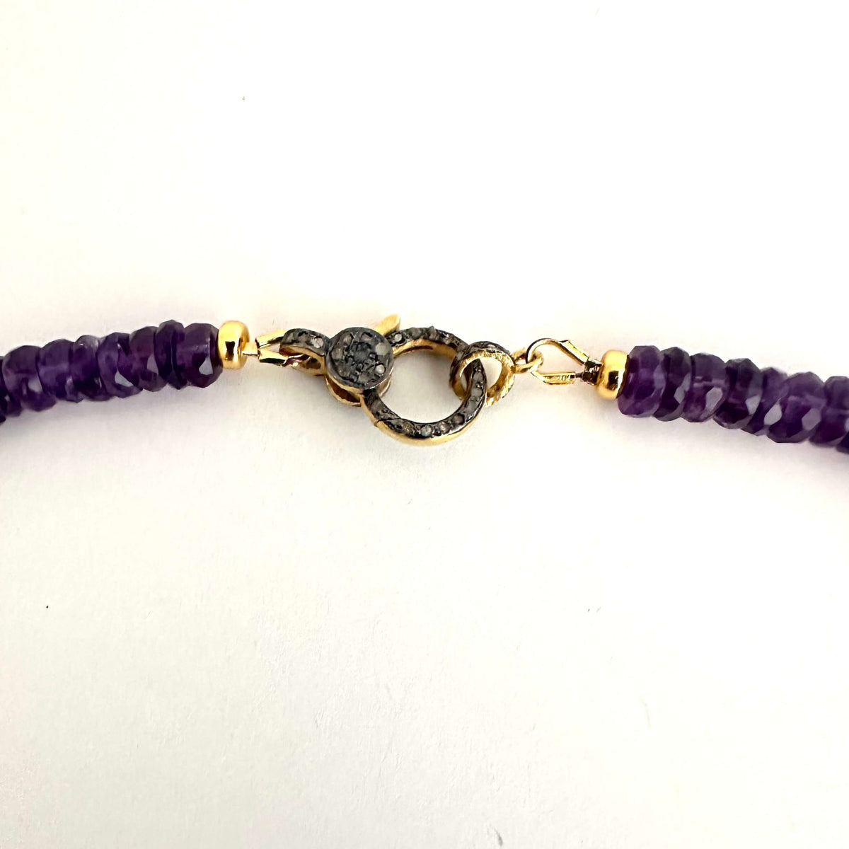 A close-up of a beaded bracelet featuring faceted purple beads and a central gold-toned clasp with a dark textured center, set against a plain white background.