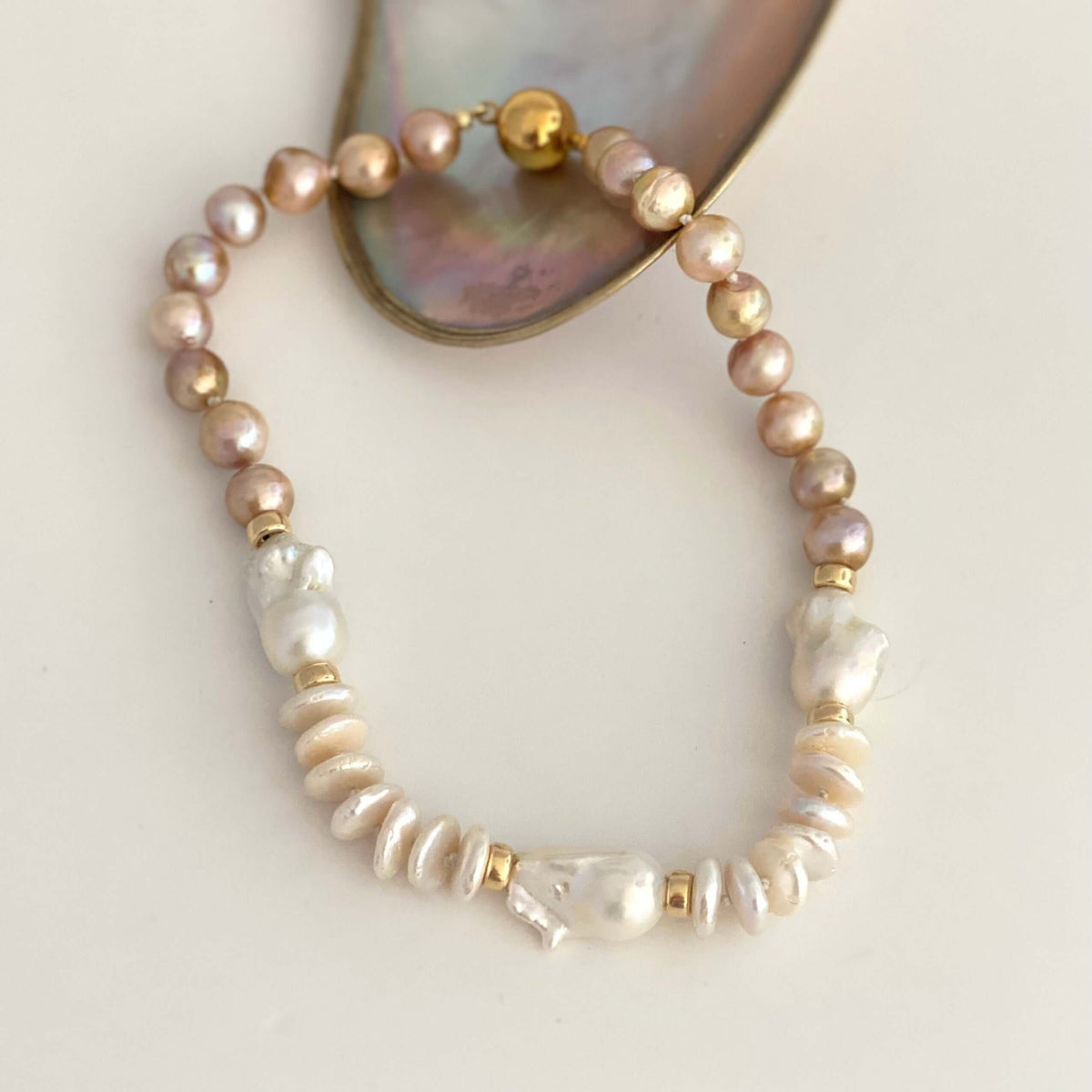 A beaded stretch bracelet featuring light pink freshwater pearls, baroque pearls, and gold spacer beads, displayed on a neutral cream background with a mother-of-pearl shell prop.