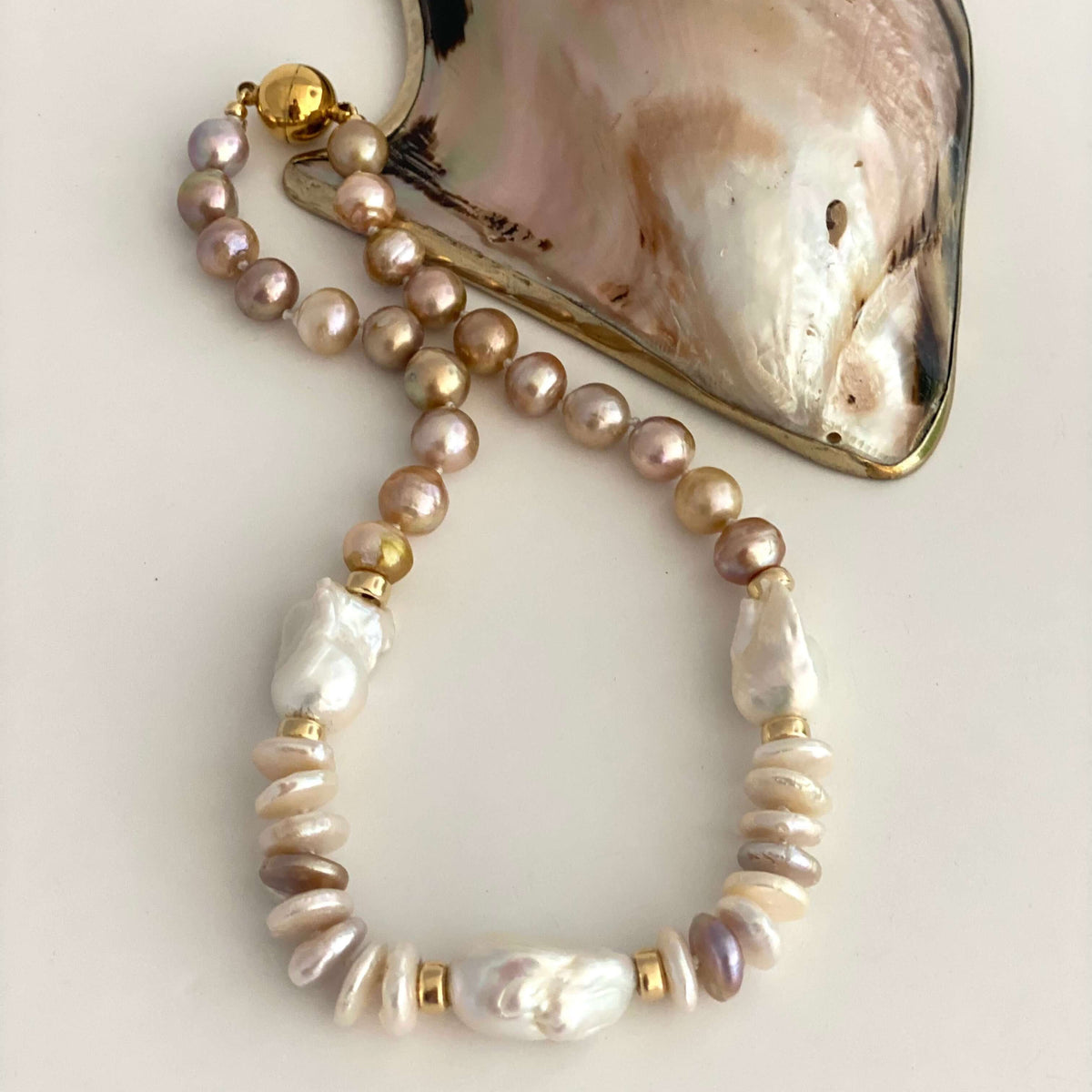 A beaded necklace featuring iridescent baroque pearls in soft pink, beige, and white, accented with textured shell beads and gold spacer rings, displayed next to a large iridescent abalone shell on a light beige background.