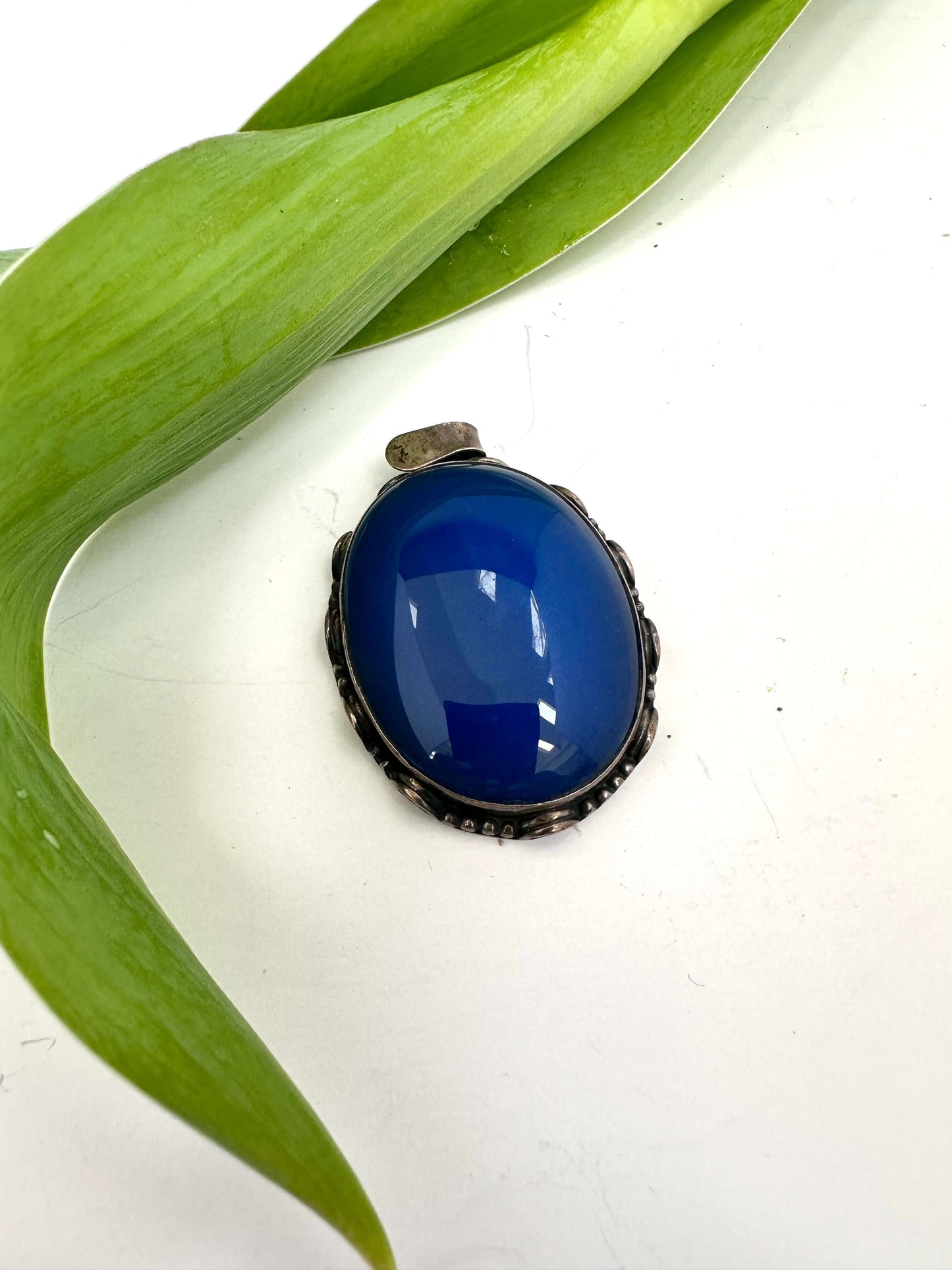 Vintage blue agate pendant in oxidized sterling silver with a deep cobalt-blue hue resting on a green leaf.