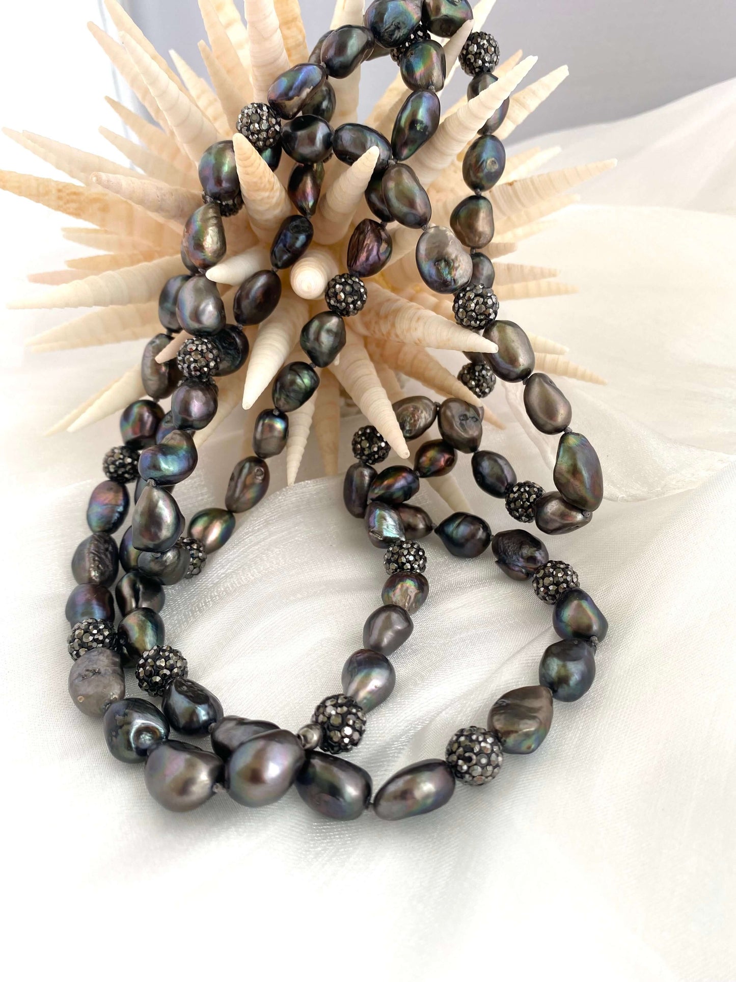Very long peacock pearl necklace features iridescent pearls and rhinestone spacer beads against a decorative background.