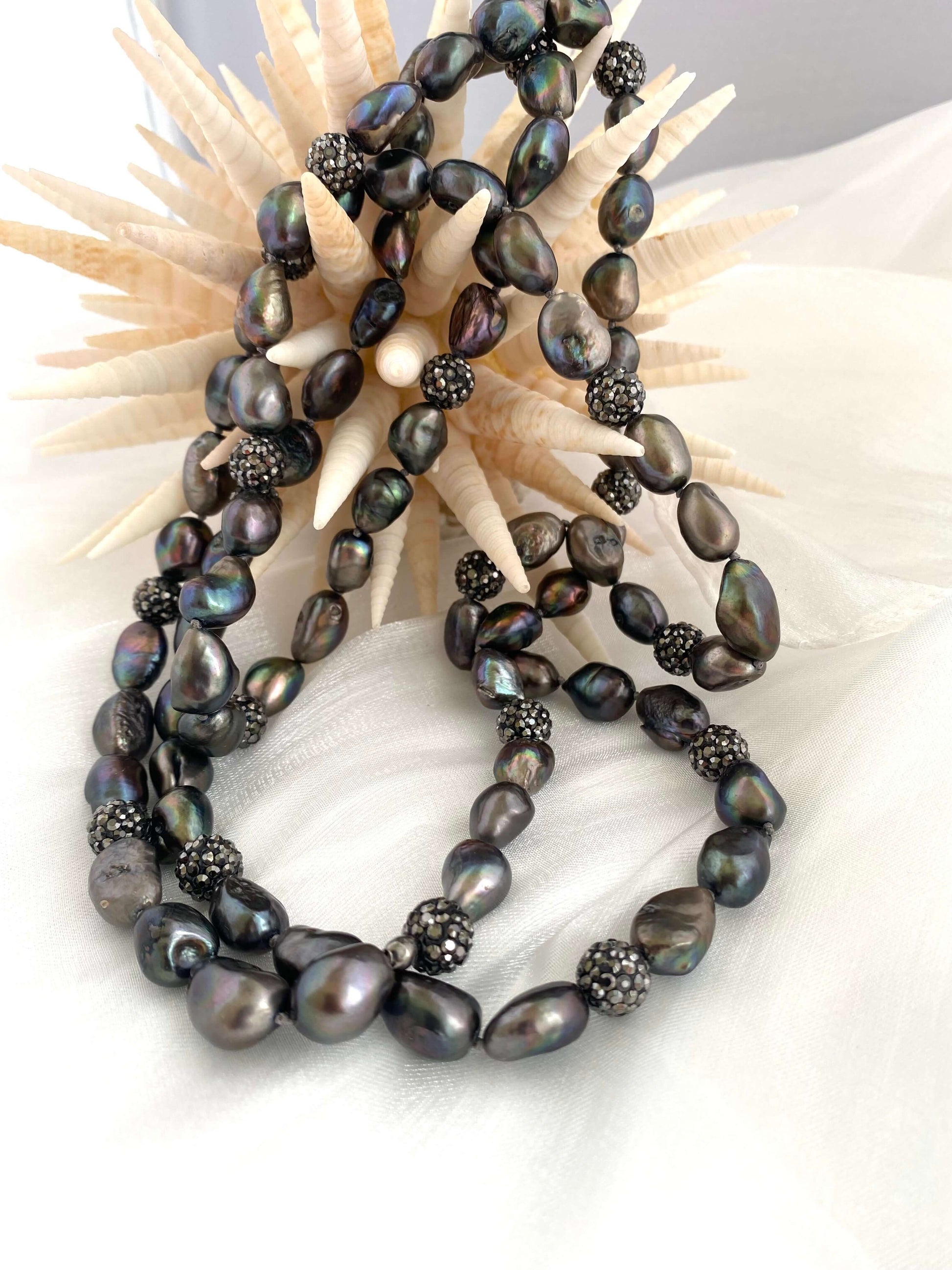 Very long peacock pearl necklace features iridescent pearls and rhinestone spacer beads against a decorative background.