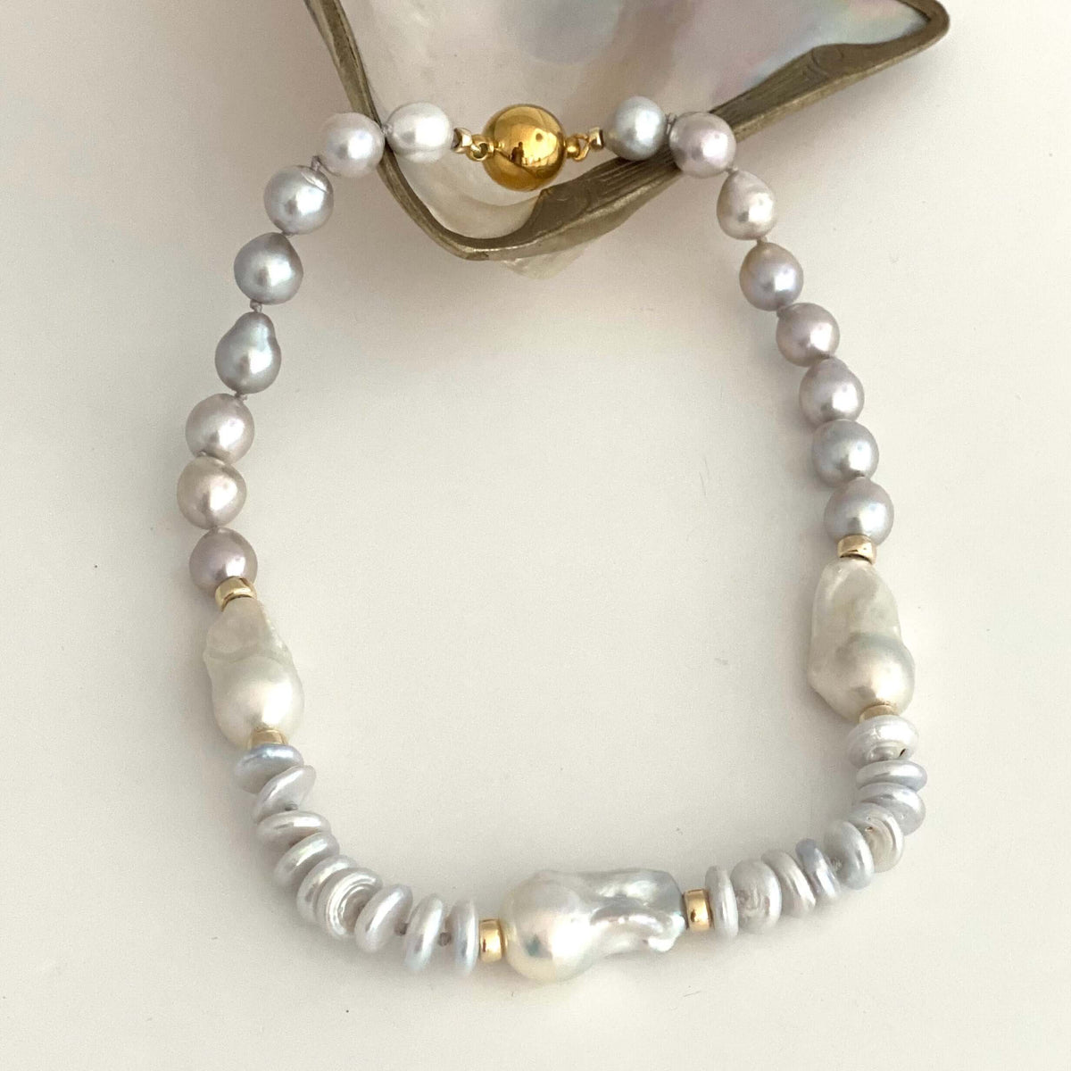 HAND KNOTTED NECKLACE made of white and silver baroque pearls, silver PEARL spacer beads, and gold bead, displayed on a white surface with a piece of mother-of-pearl shell at the top.