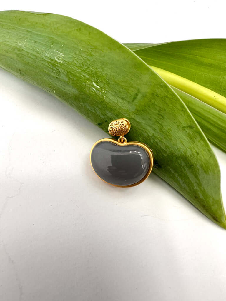 Hotan jade charm pendant in gold bezel showcasing smooth gray nephrite jade on a green leaf background.