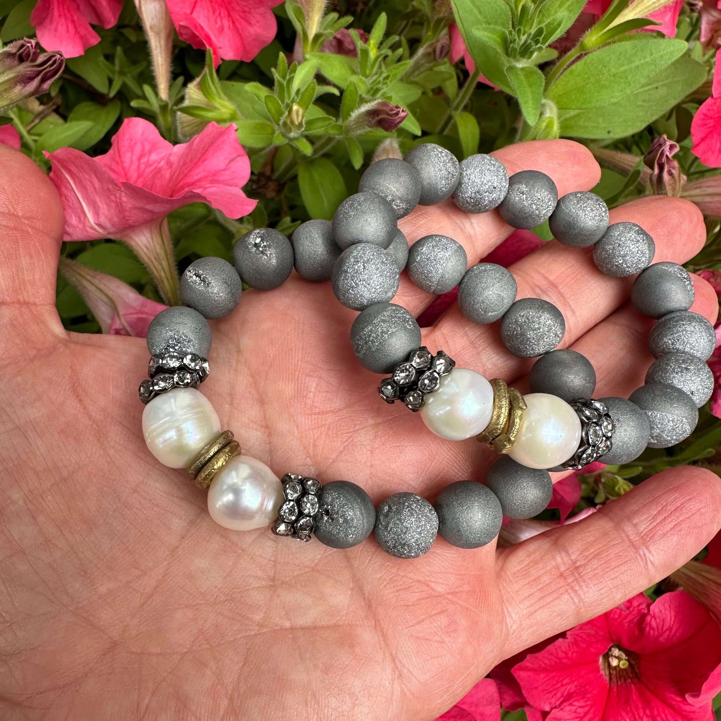 Two grey druzy agate and pearl bracelets held in hand against pink flowers