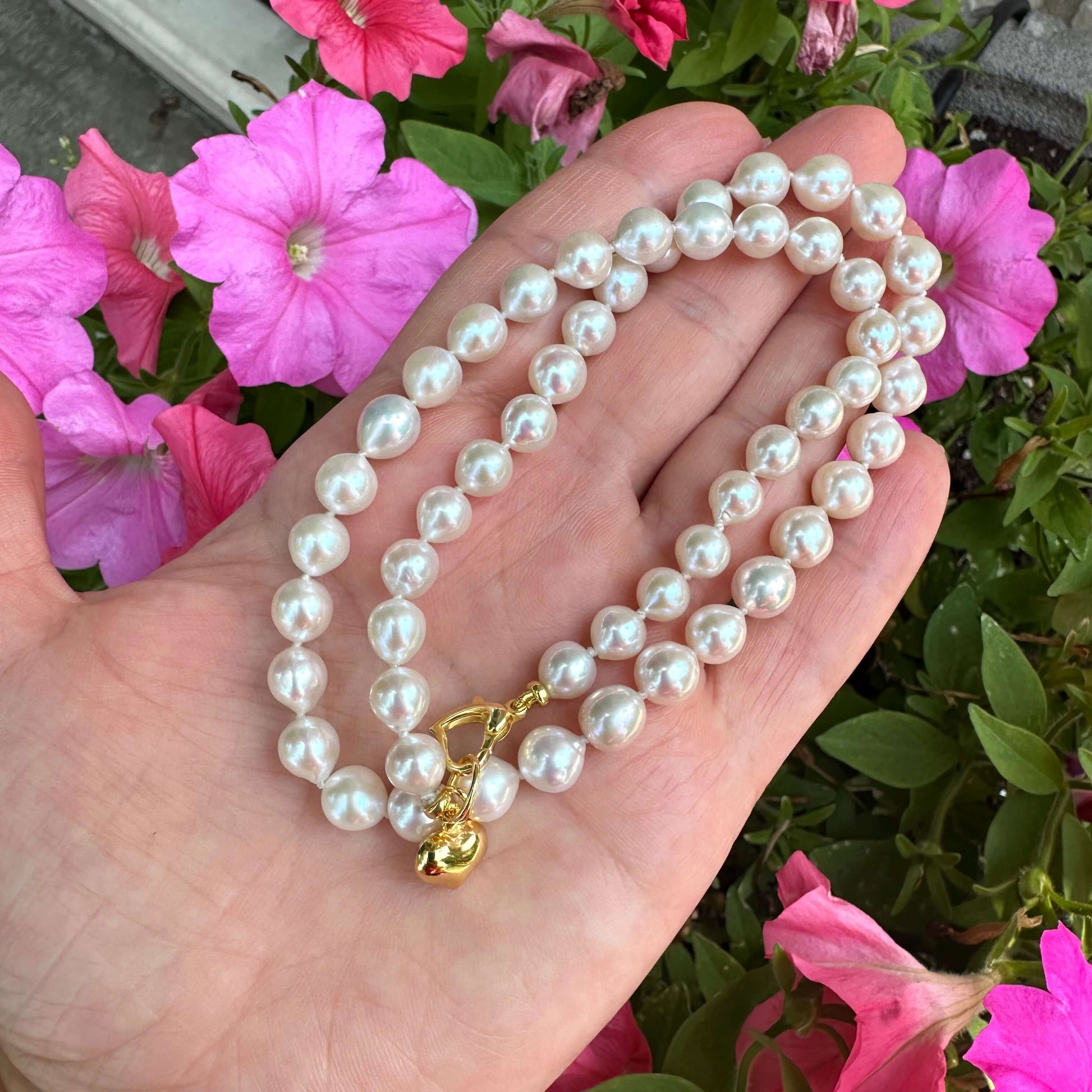 Classic pearl necklace with gold heart charm held in hand, showcasing lustrous pearls and vibrant flowers in background.