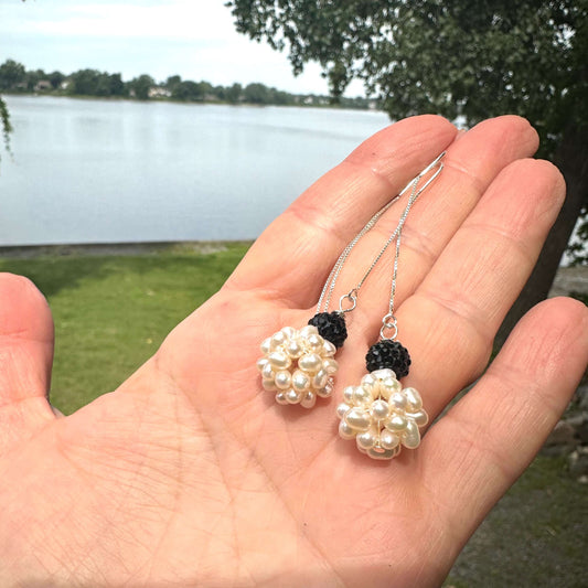 White pearl threader earrings with black rhinestones held in hand against green foliage