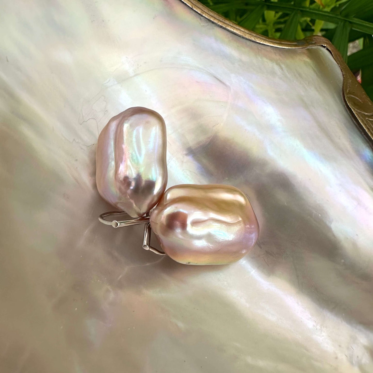 Purple pearl drop earrings displayed on pearl shell with green leaves, showing natural color variation and elegant silver hooks.