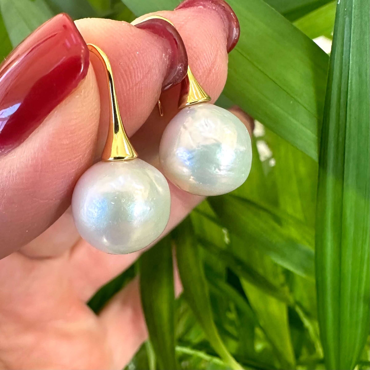 Hand holding white pearl drop earrings outdoors, showing large Edison pearls and gold vermeil hooks under natural daylight.