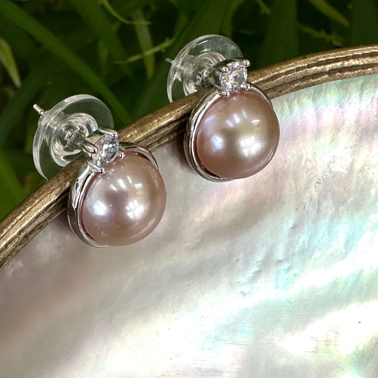 Handcrafted pink mabe pearl stud earrings resting on white shell with soft greenery, showing luminous blush hue and silver setting.