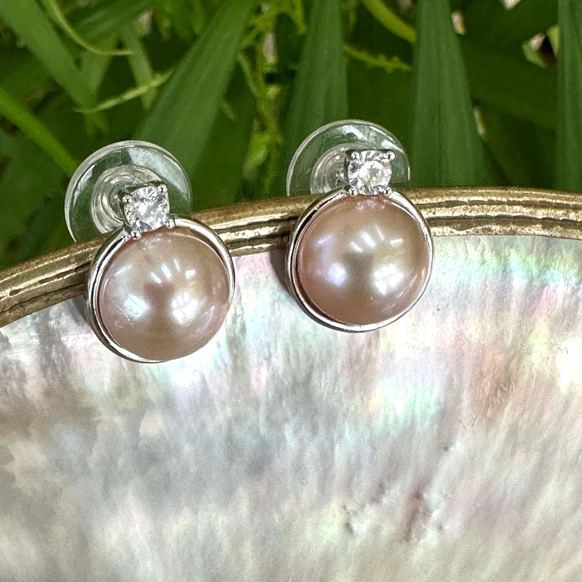Pink mabe pearl stud earrings with CZ in sterling silver, close-up dangling from white pearl shell with green foliage background.