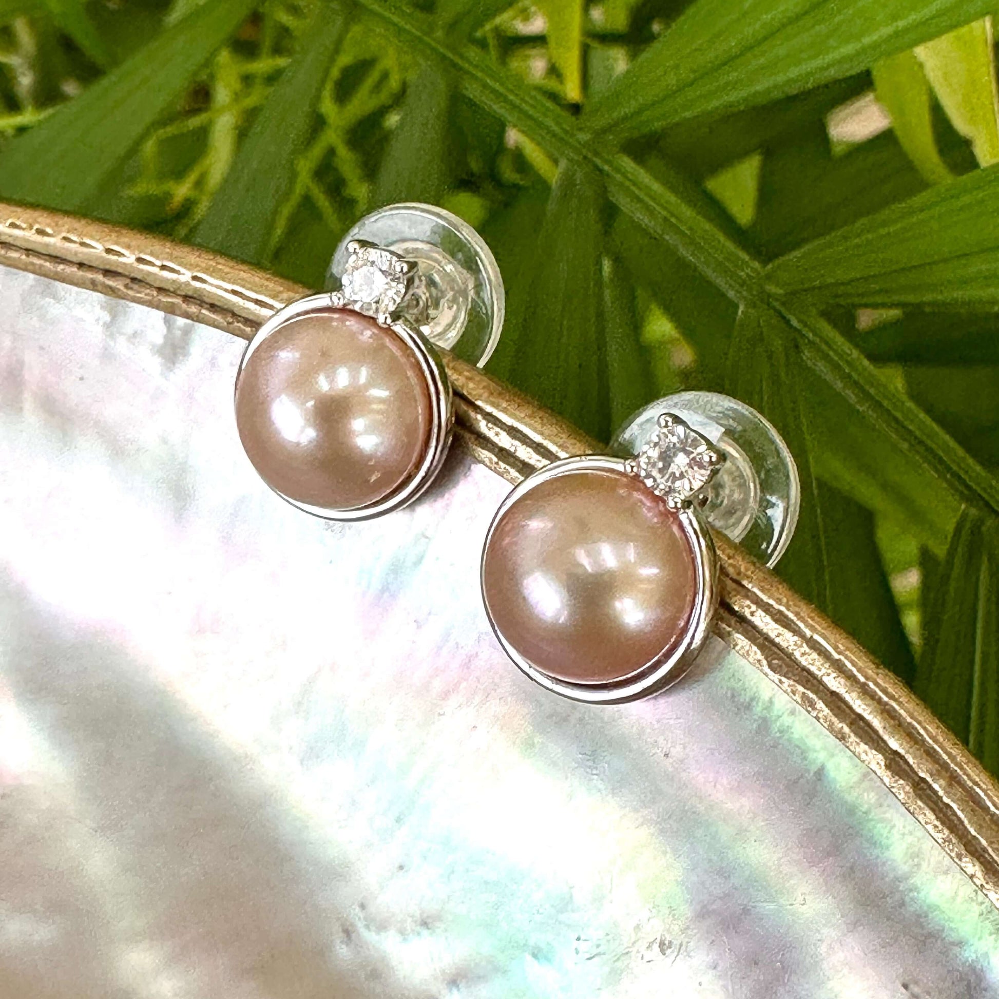 Elegant pink mabe pearl stud earrings displayed on white pearl shell among green leaves, highlighting natural luster and CZ sparkle.