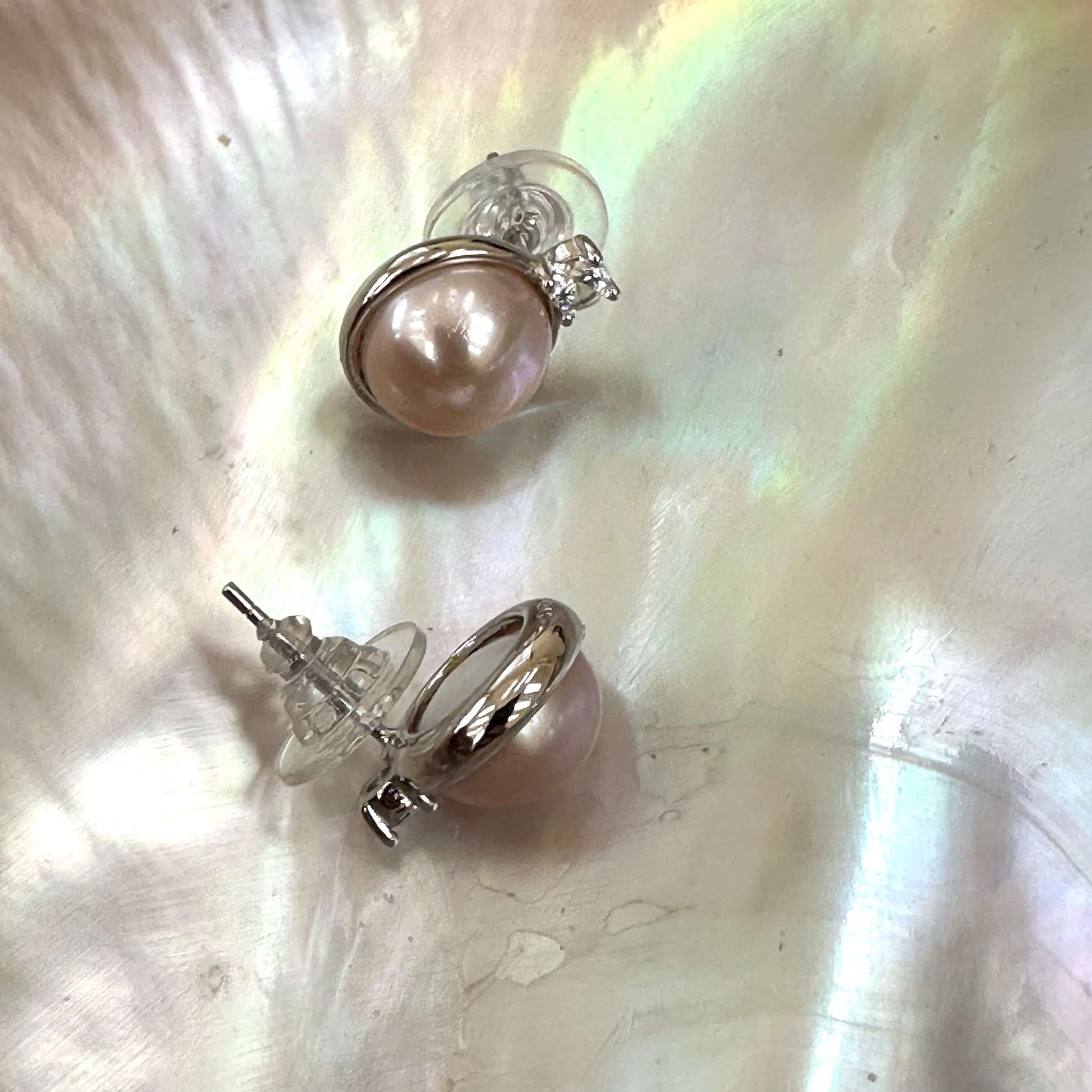 Detailed view of pink mabe pearl stud earrings on white shell, capturing silver bezel setting and soft pastel glow of pearls.