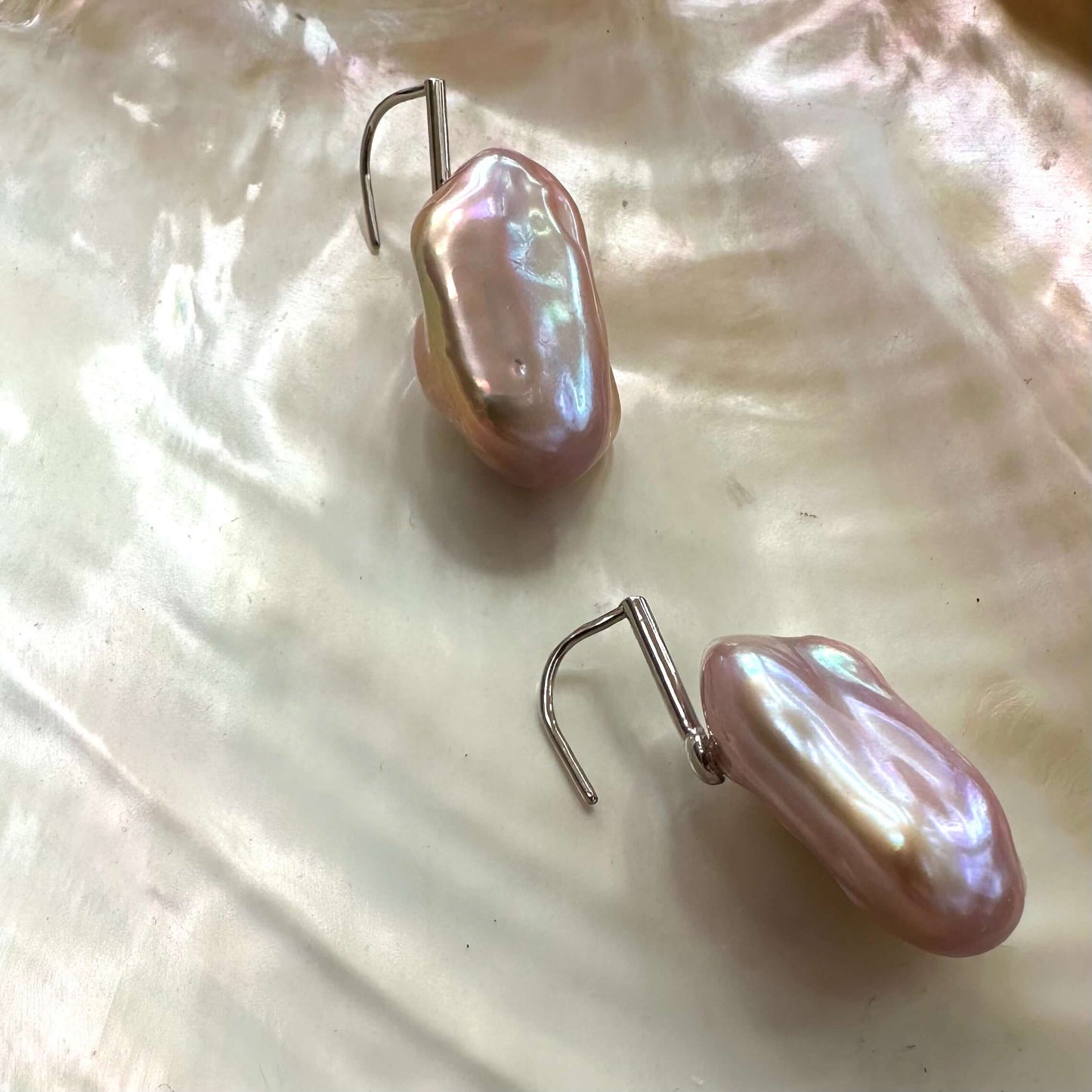 Rectangular baroque purple pearl drop earrings resting on white shell, showing unique shape and shimmering multitone surface.