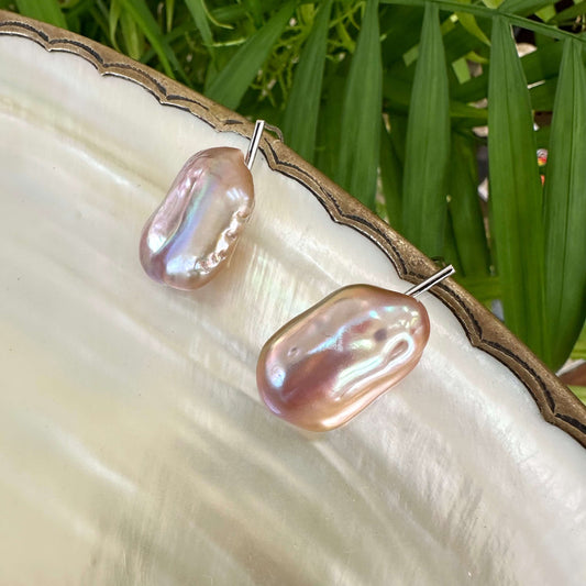 Rectangular purple pearl drop earrings on shell among greenery, highlighting iridescent tones and handcrafted sterling silver finish.