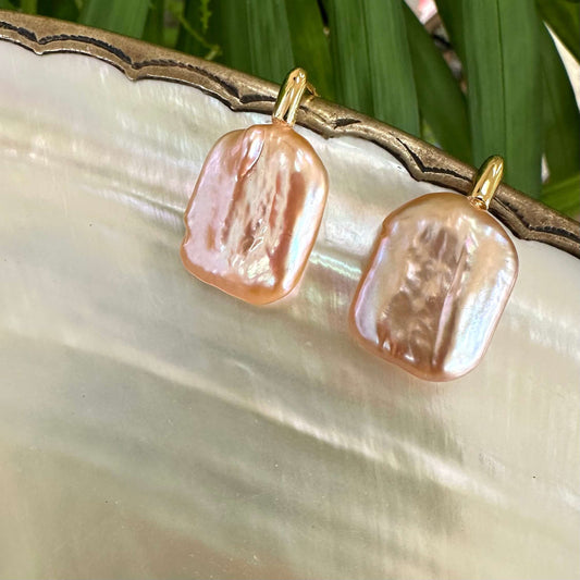 Golden peach pearl drop earrings on shell among foliage, highlighting radiant peach color and artistic baroque pearl detail.