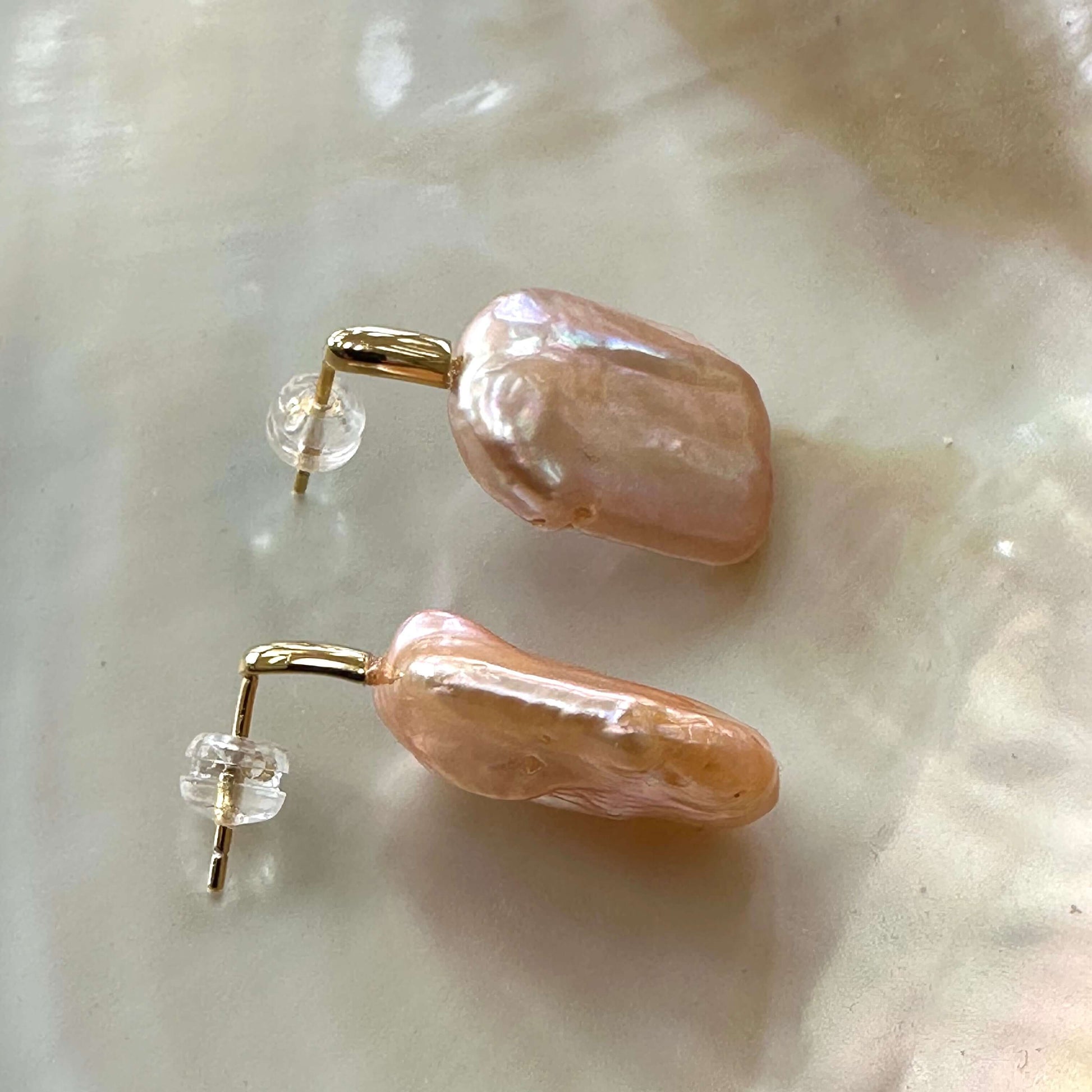 Handmade golden peach pearl drop earrings resting on white pearl shell, highlighting warm iridescent glow and gold vermeil posts.