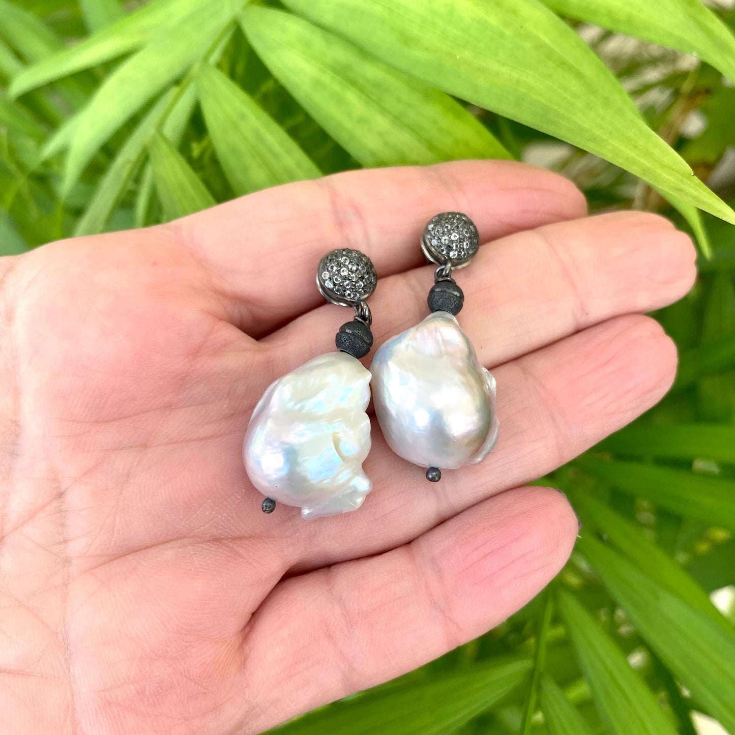 Baroque pearl drop earrings featuring diamond pavé studs in oxidized silver, displayed in a hand with green leaves.