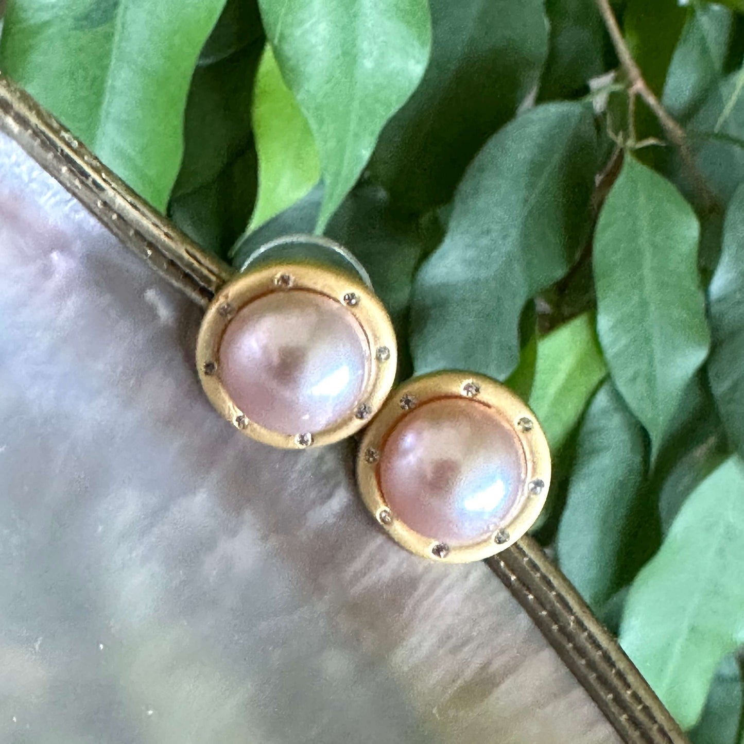 Pink mabe pearl stud earrings with CZ on white shell and green leaves, showing natural light reflection and organic charm.