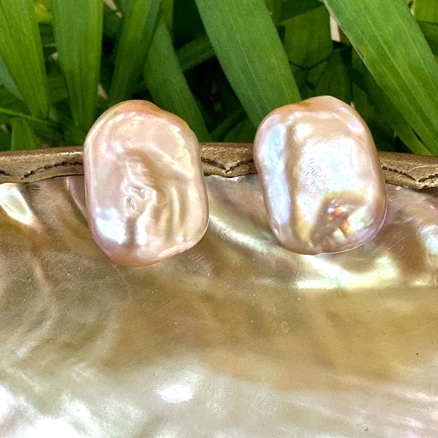 Golden pink rectangle baroque pearl stud earrings hanging from white pearl shell with green leaf background, designed and handmade in Canada.
