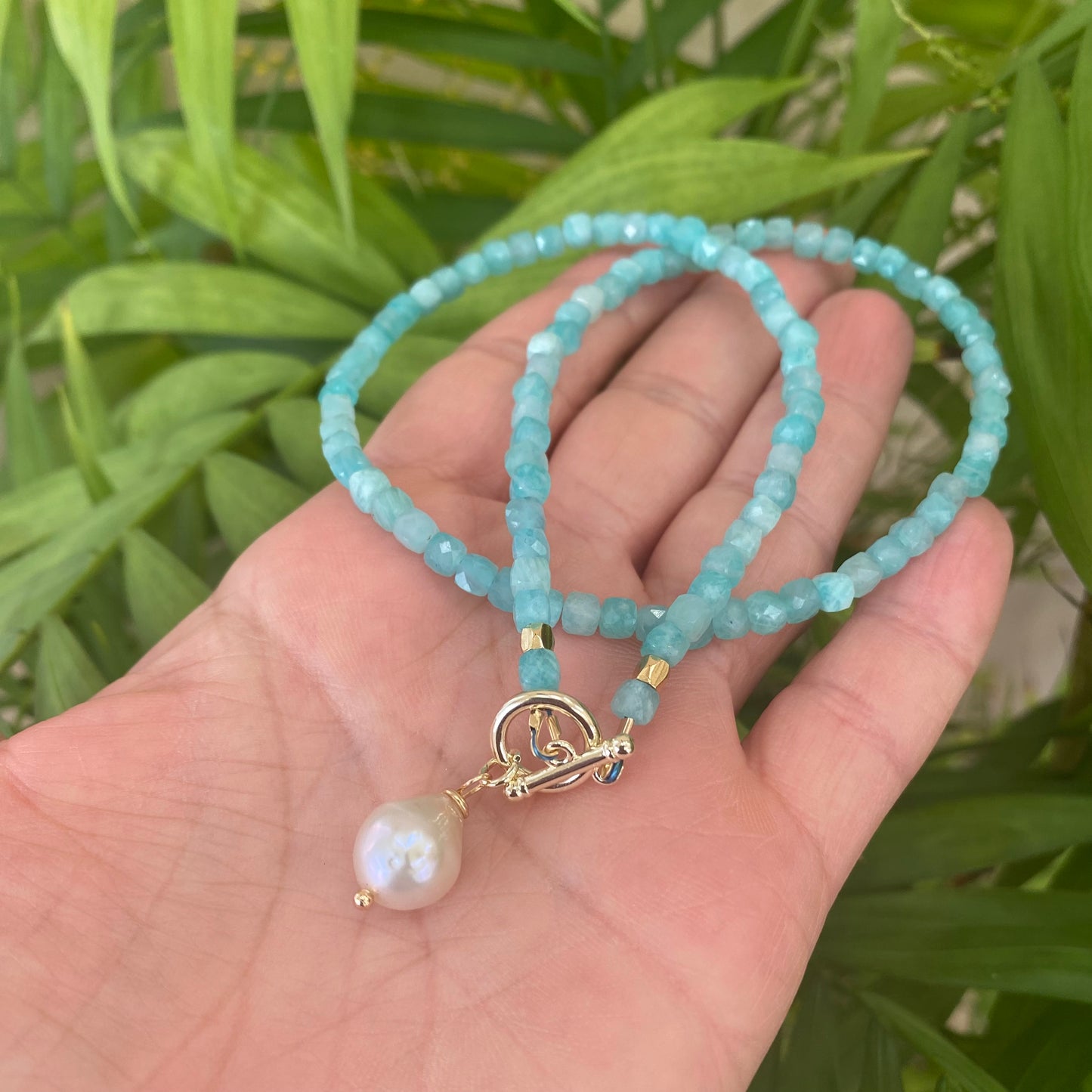Blue Amazonite Baroque Pearl Necklace | Toggle & Charm Design displayed in a hand with lush green background.
