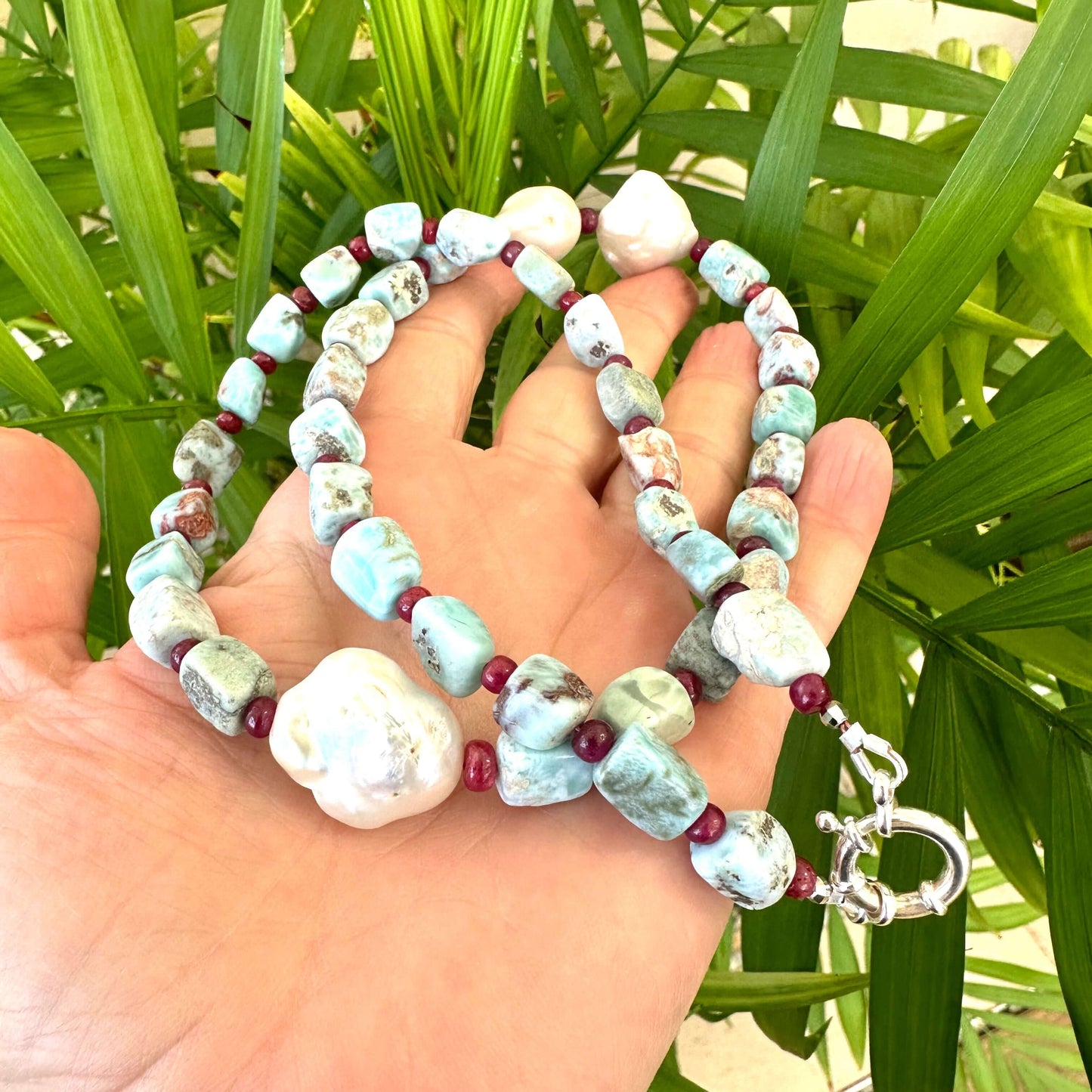 Hand holding larimar ruby and baroque pearl necklace near green leaf background