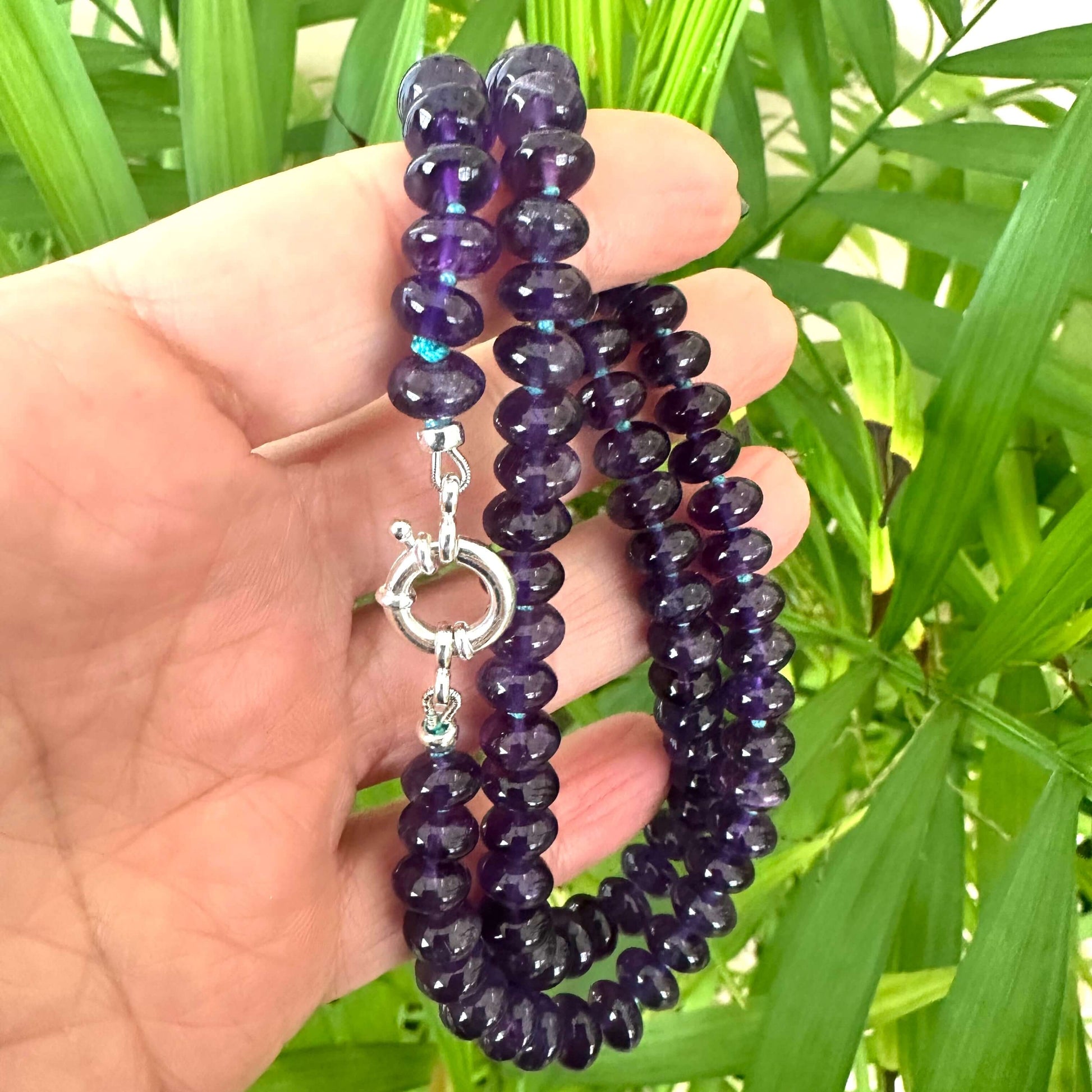 Amethyst beaded necklace held in hand for size, against green leaf background