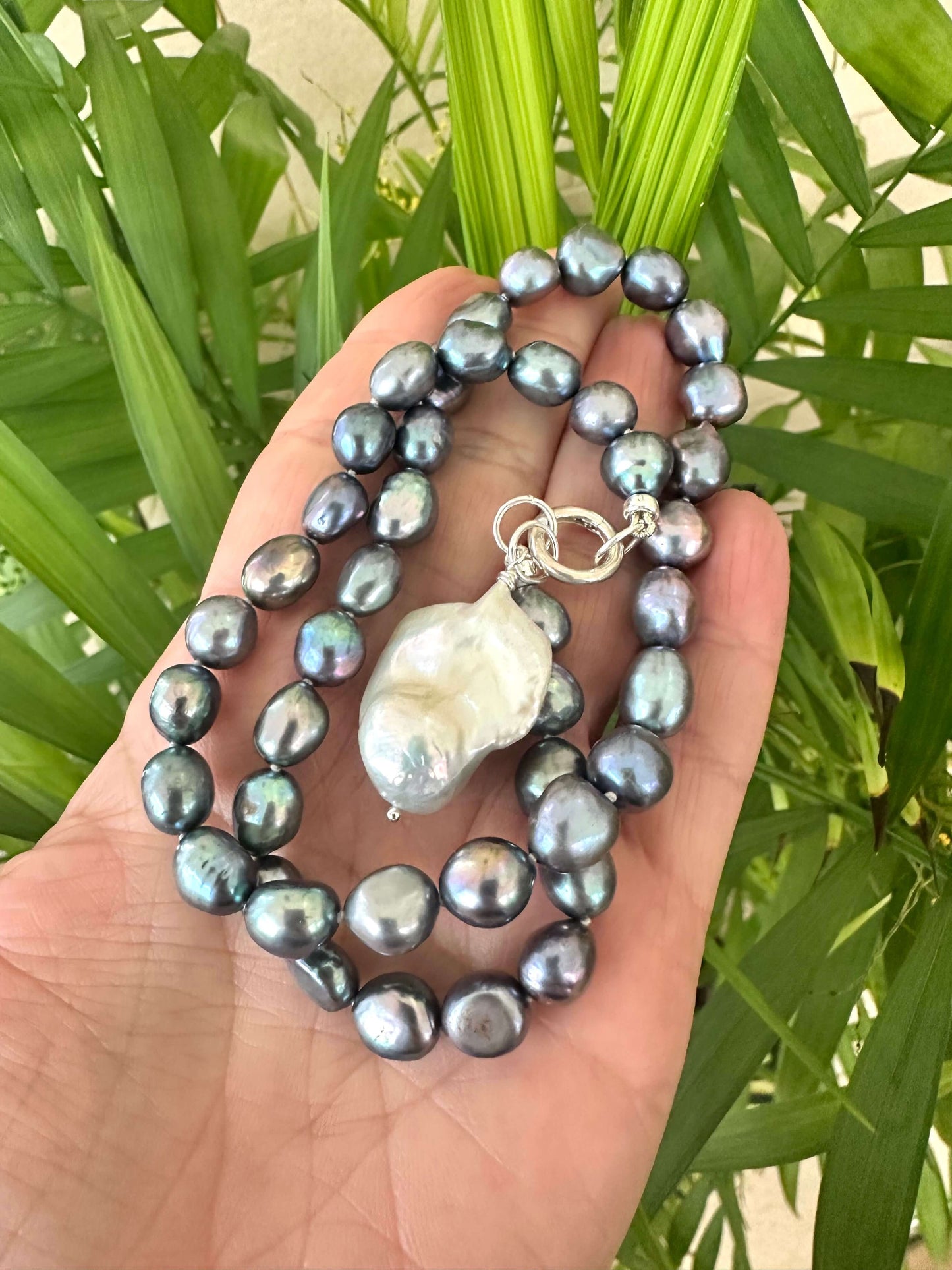 Hand holding black pearl necklace with baroque pendant near green leaf background