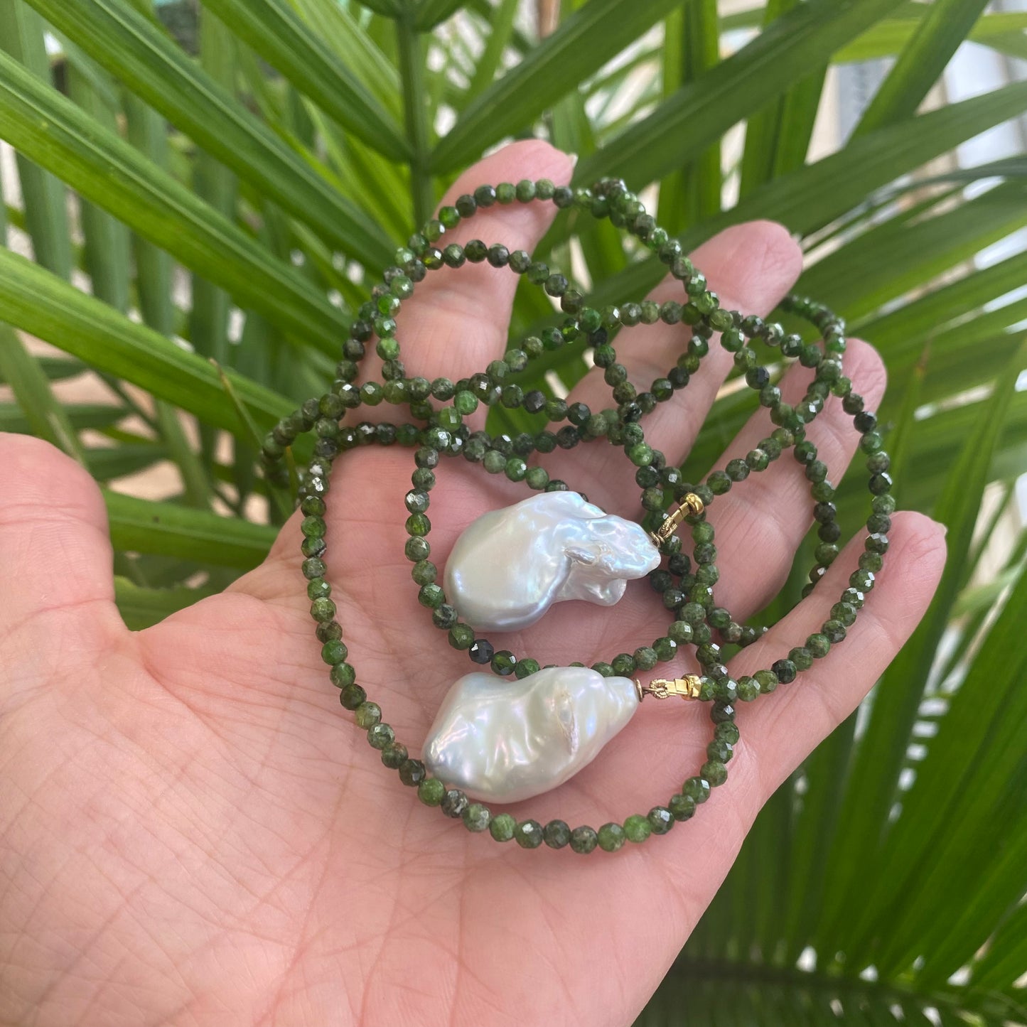 Green diopside lariat necklace with Baroque pearls displayed in a hand surrounded by green foliage.