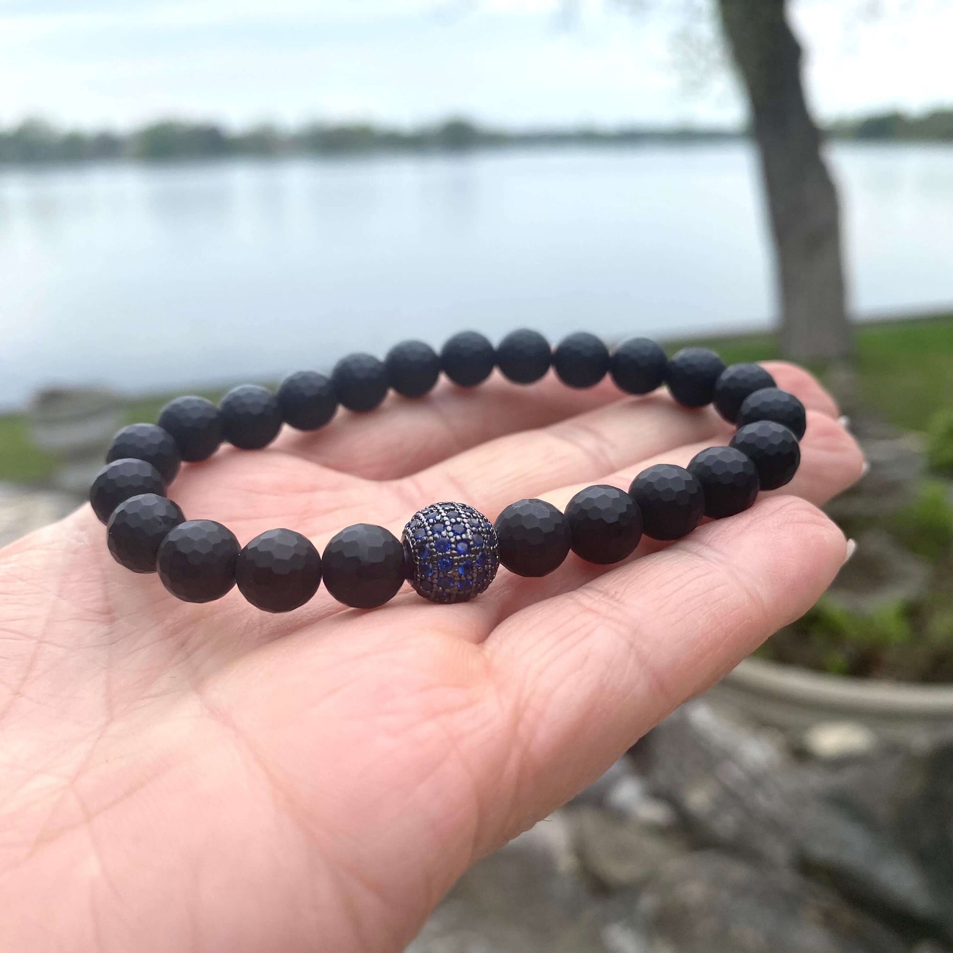Matte black onyx mens bracelet with blue CZ centerpiece, shown in hand against a serene lake background.