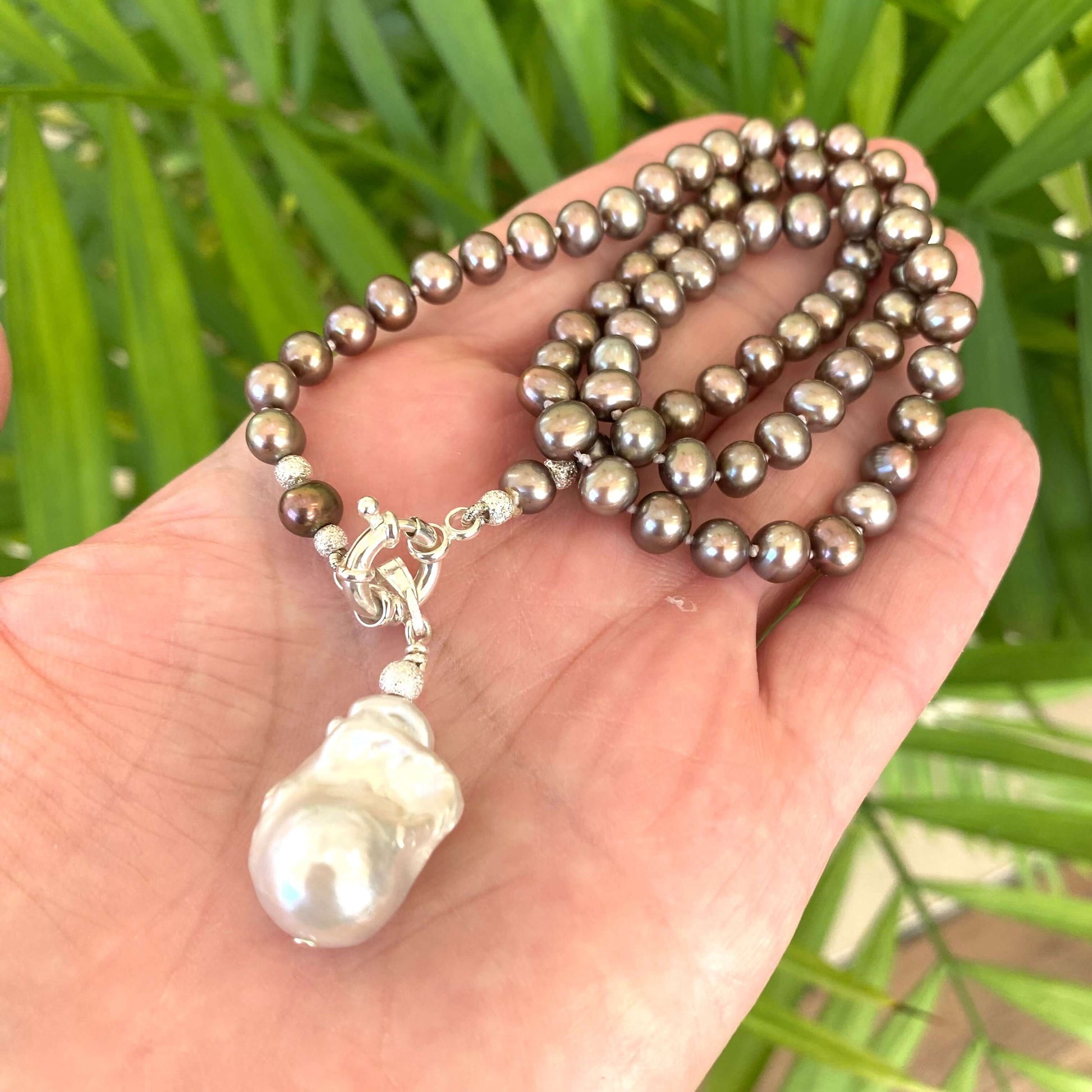 Hand holding bronze pearl necklace with removable white pendant against green leaves, showing iridescence and organic form.