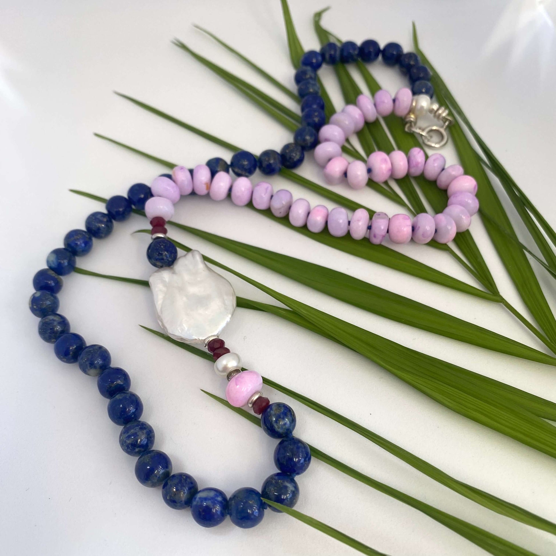 Close-up of lapis lazuli and baroque pearl necklace with ruby detail on white and leaf background.