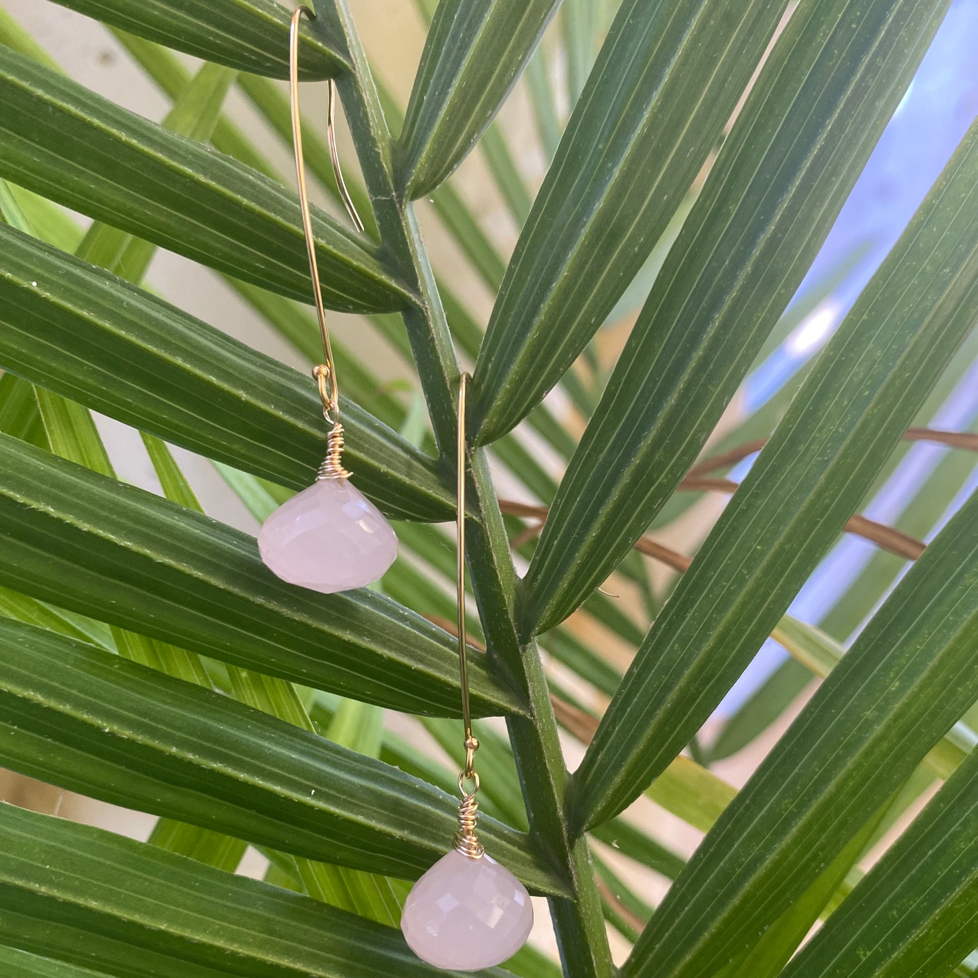 Rose quartz threader earrings with briolettes hanging on a green palm leaf, showcasing their delicate design.