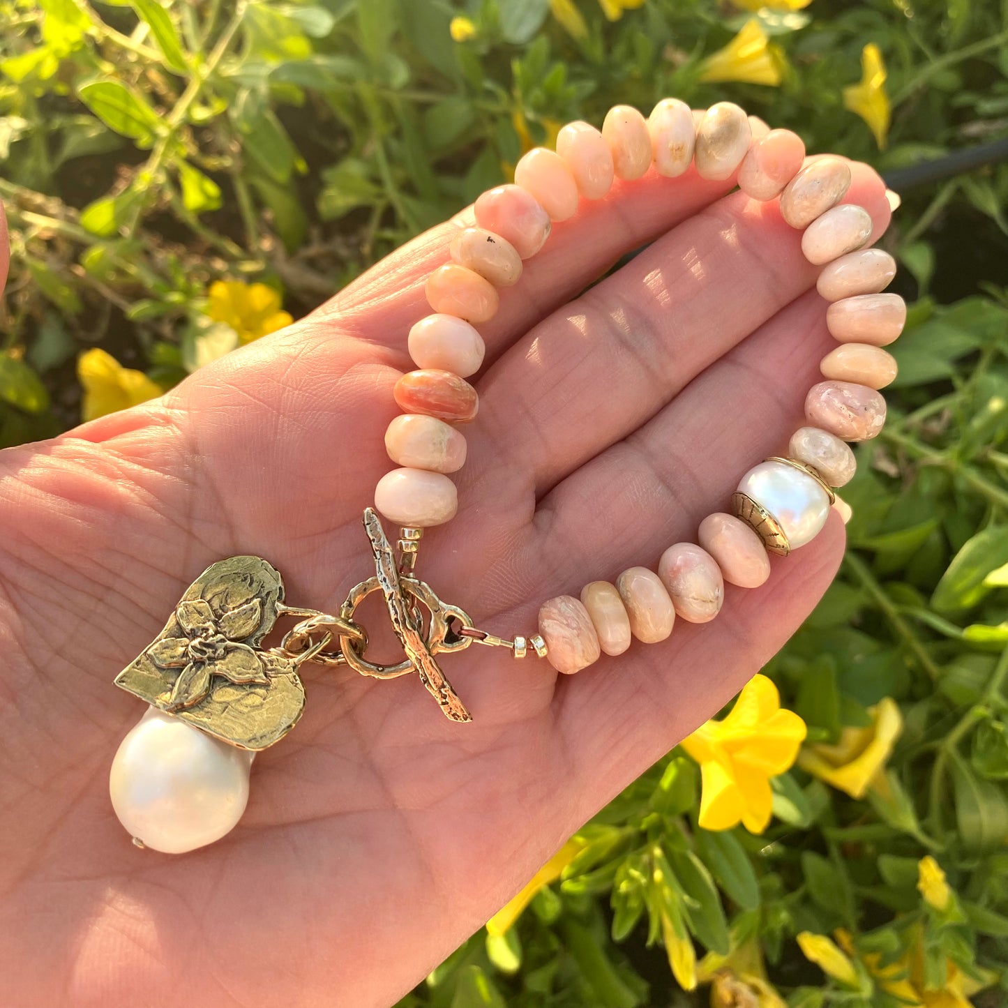 Pink opal bracelet with heart and baroque pearl charm displayed on hand, highlighting its artisan craftsmanship.