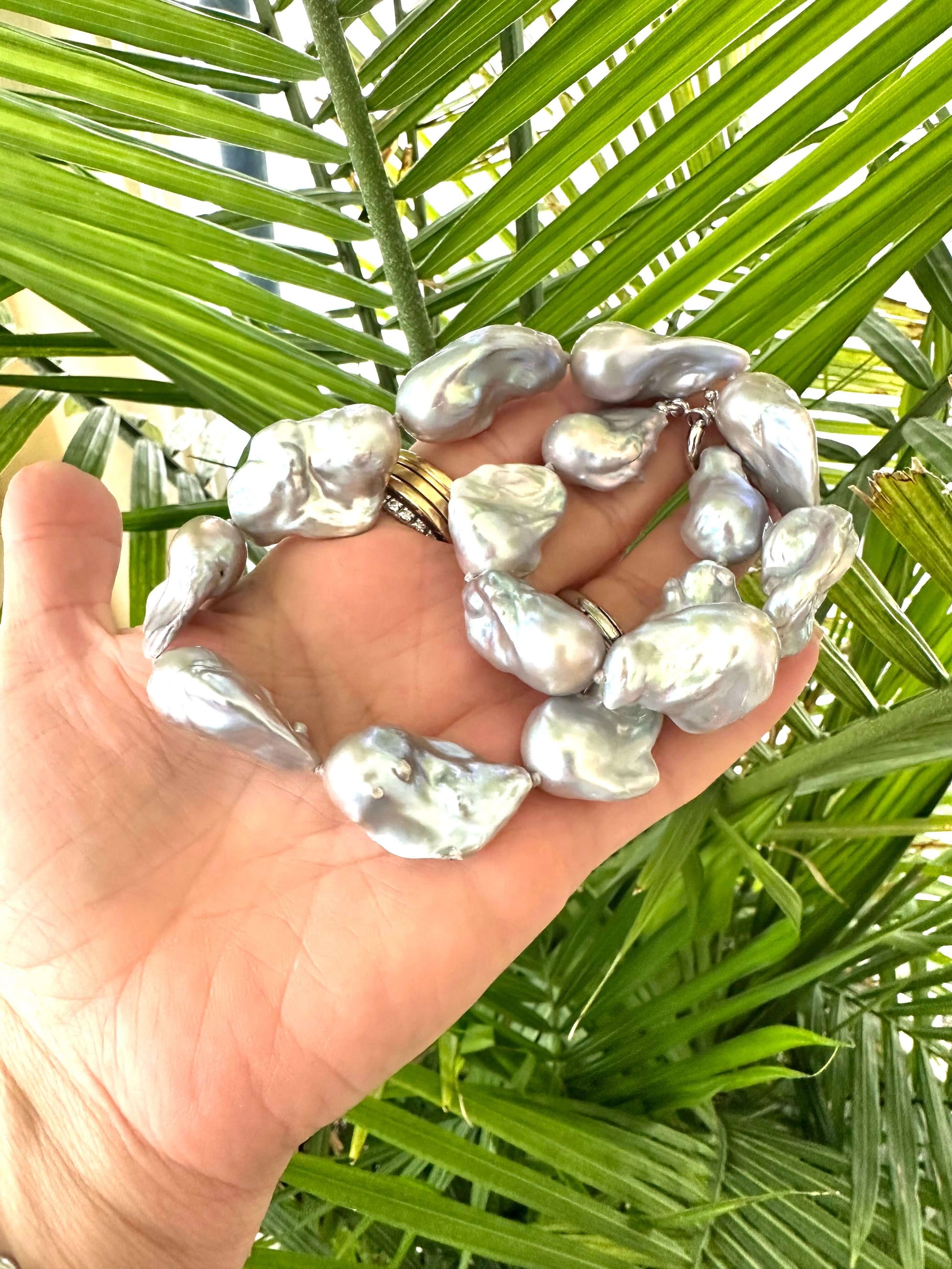 Hand holding a baroque pearl necklace with silver details, showcasing large, iridescent pearls against a green background.