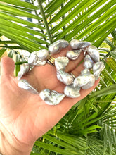 Charger l&#39;image dans la galerie, Hand holding a baroque pearl necklace with silver details, showcasing large, iridescent pearls against a green background.
