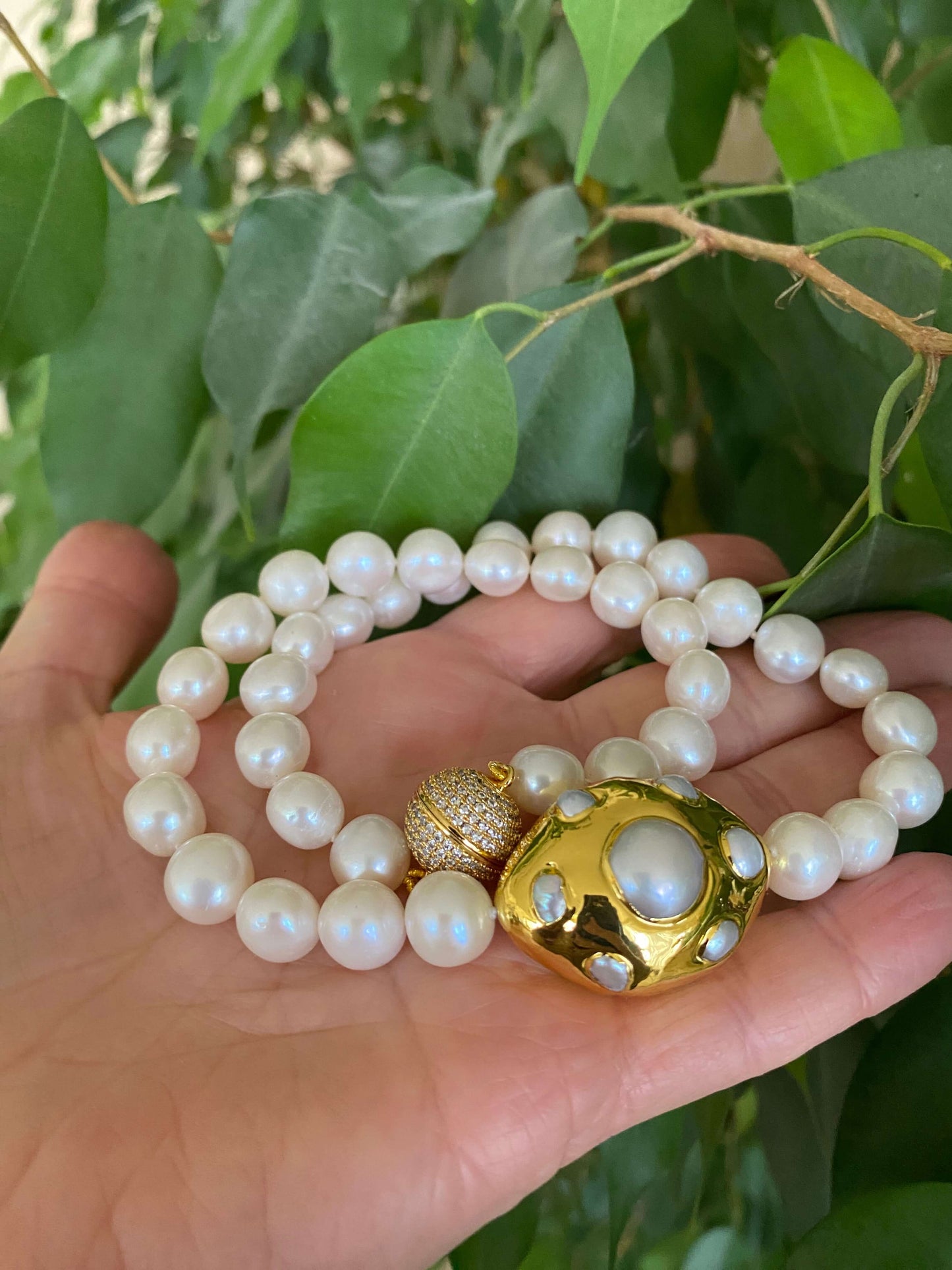 White potato pearl necklace with gold focal accent displayed on a hand surrounded by green leaves.
