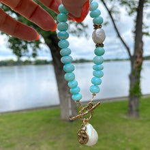 Load image into Gallery viewer, Sky blue opal bracelet with baroque pearls and cross charm, captured by a lakeside setting.
