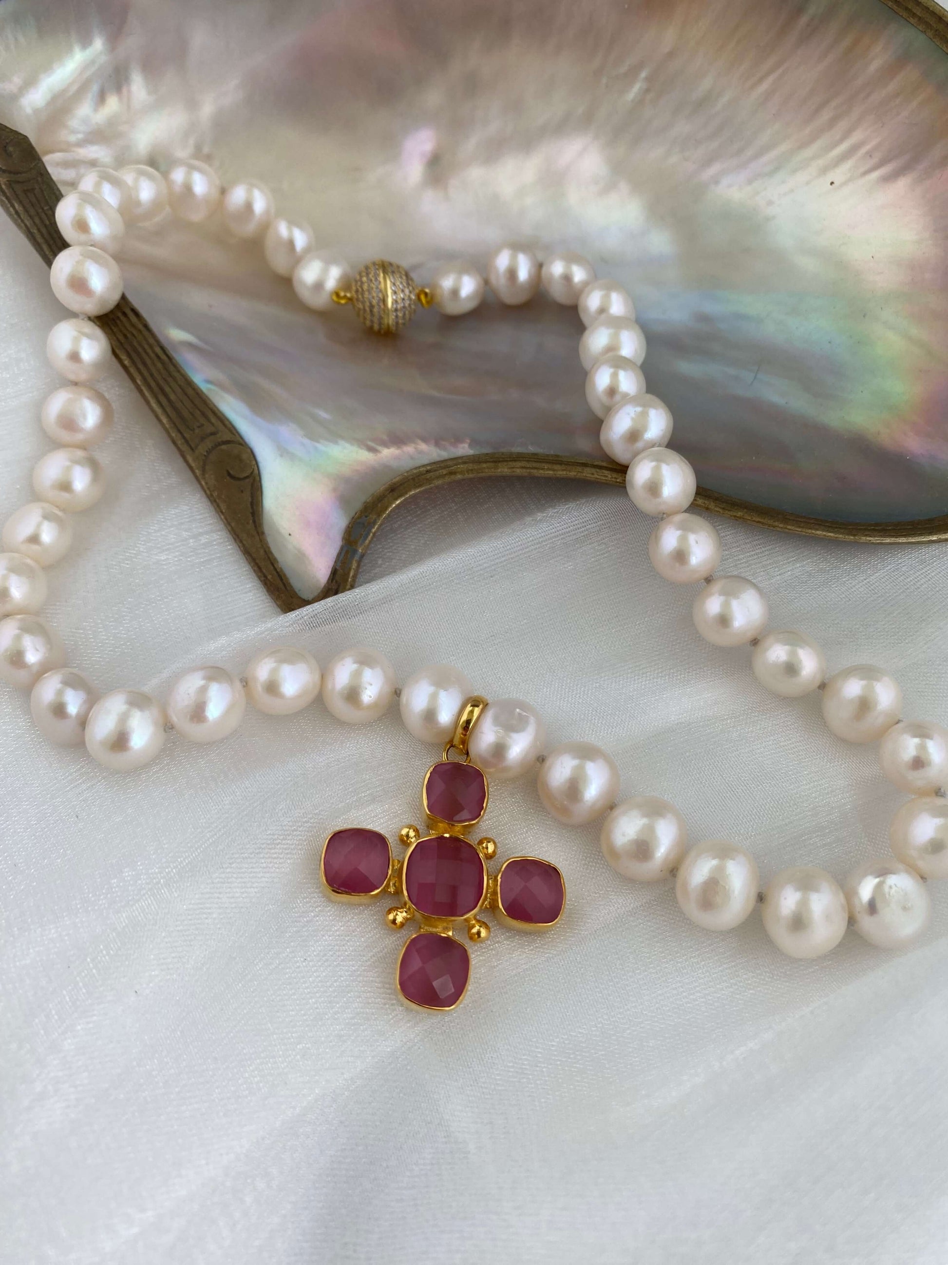 White potato pearl necklace with cross pendant featuring pink stones and gold accents, laid on an iridescent shell.