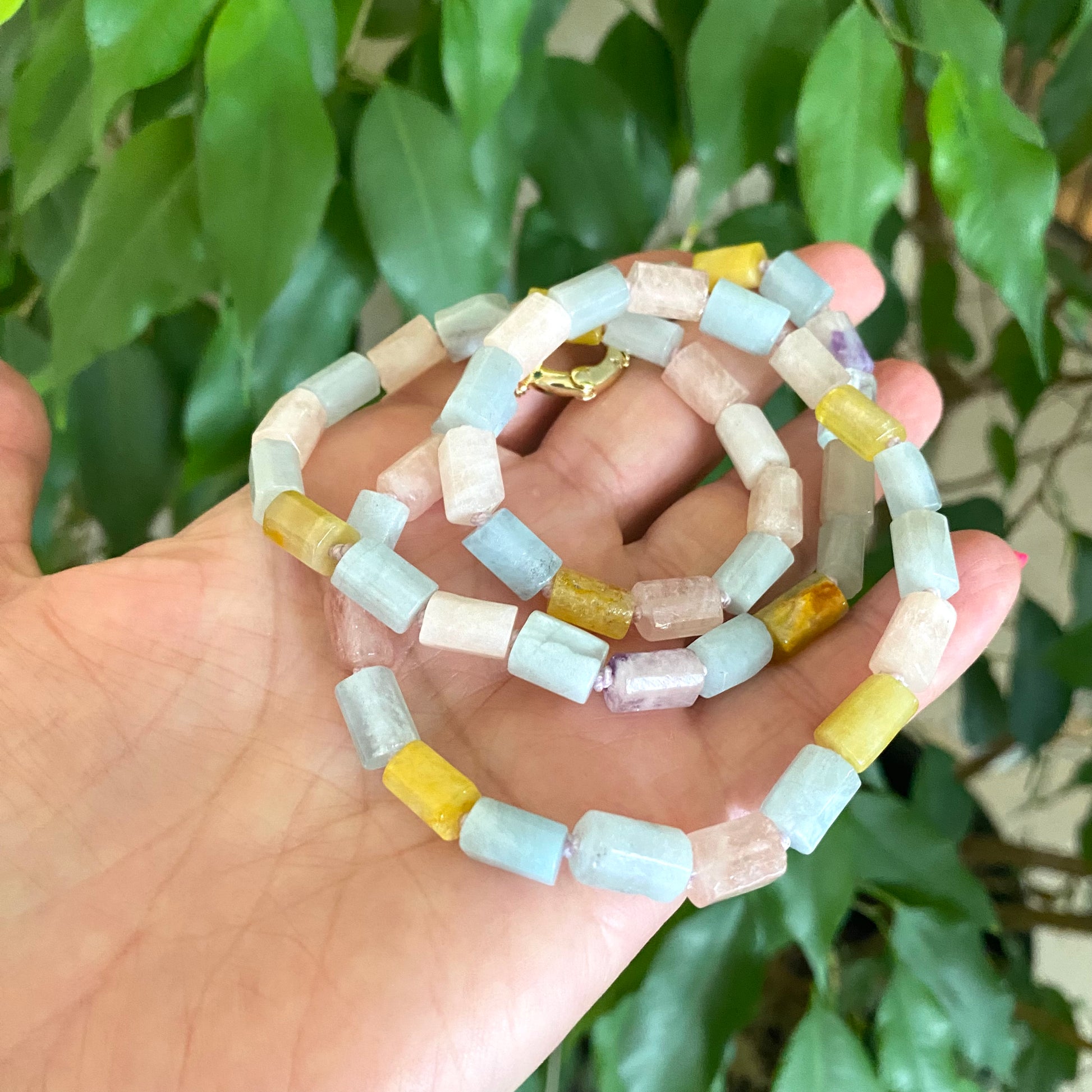 Beryl gemstone necklace featuring aquamarine, morganite, heliodor, and goshenite on a hand against green foliage.