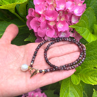 garnet toggle necklace with baroque pearl charm held in hand, surrounded by blooming pink hydrangeas