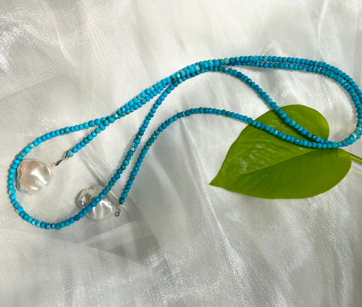 Turquoise lariat necklace with two baroque pearls on a soft fabric background and a green leaf.