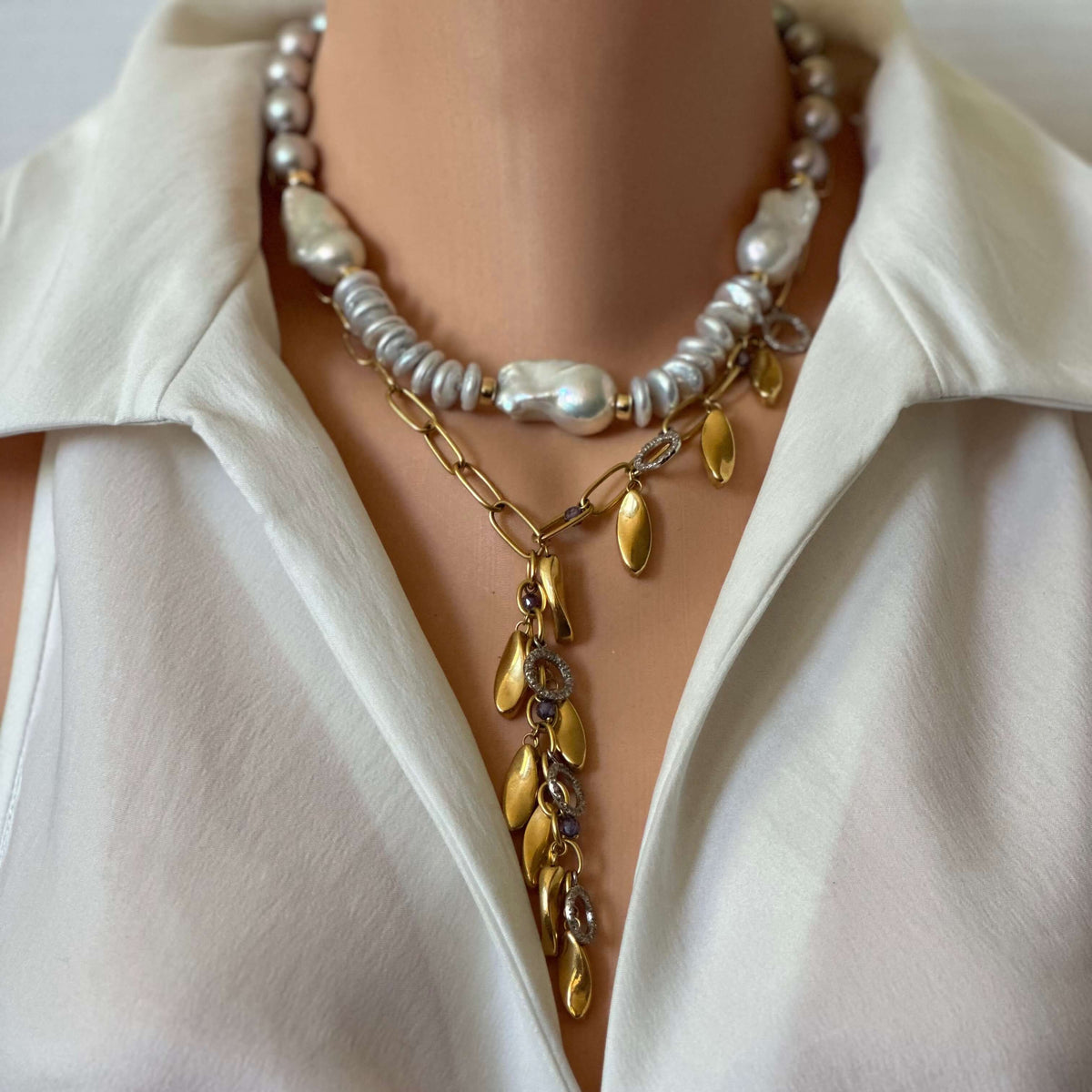 A close-up of a person wearing a layered necklace set over a white collared blouse. The necklace features a top layer with white and silver baroque pearls, a middle layer with a gold chain and gold leaf-shaped charms, and a bottom layer with a gold chain and gold leaf charms.