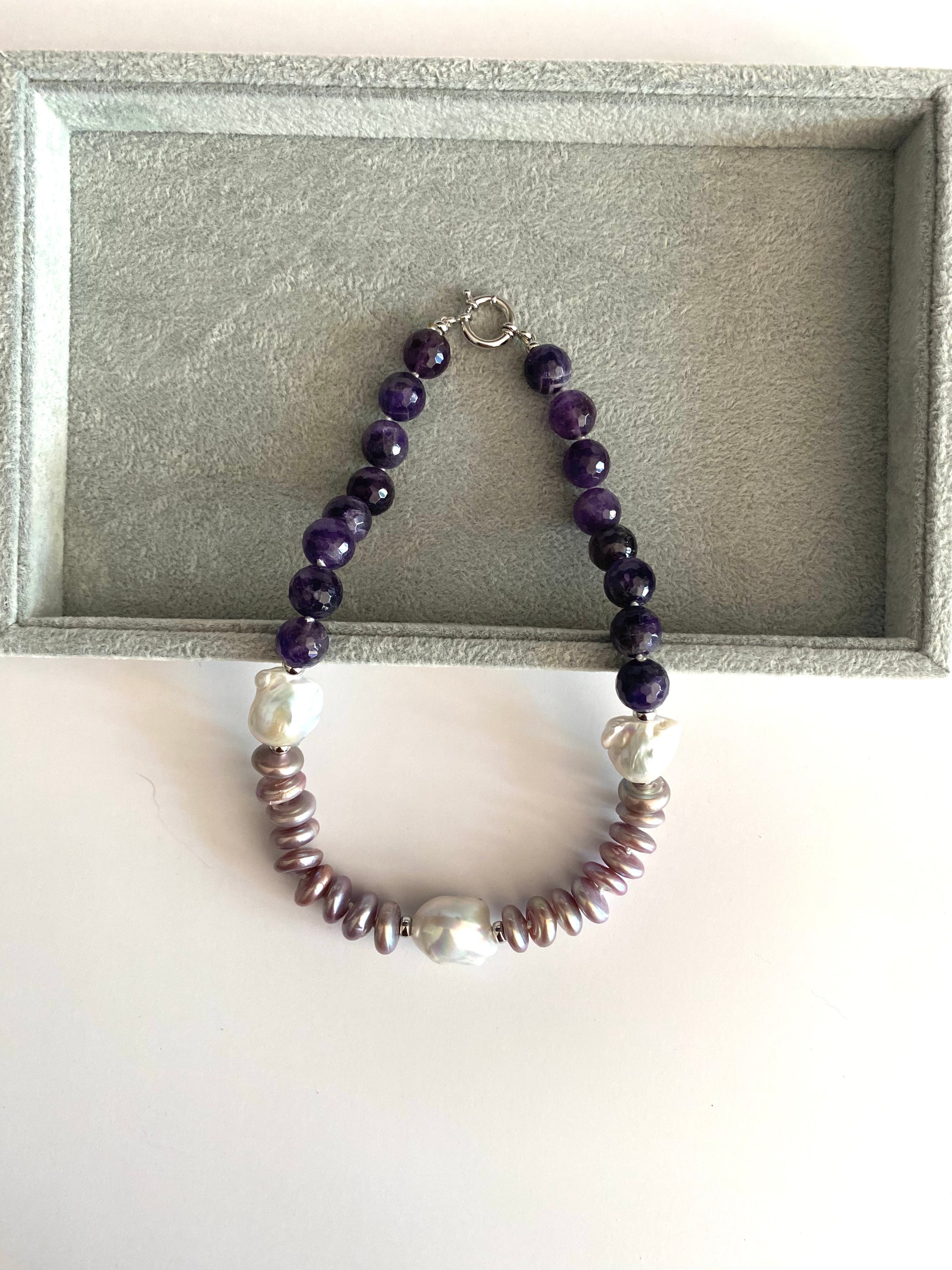 A beaded necklace with dark purple round beads, white pearl-like beads, and metallic bronze disc beads, displayed on a gray velvet jewelry box insert.